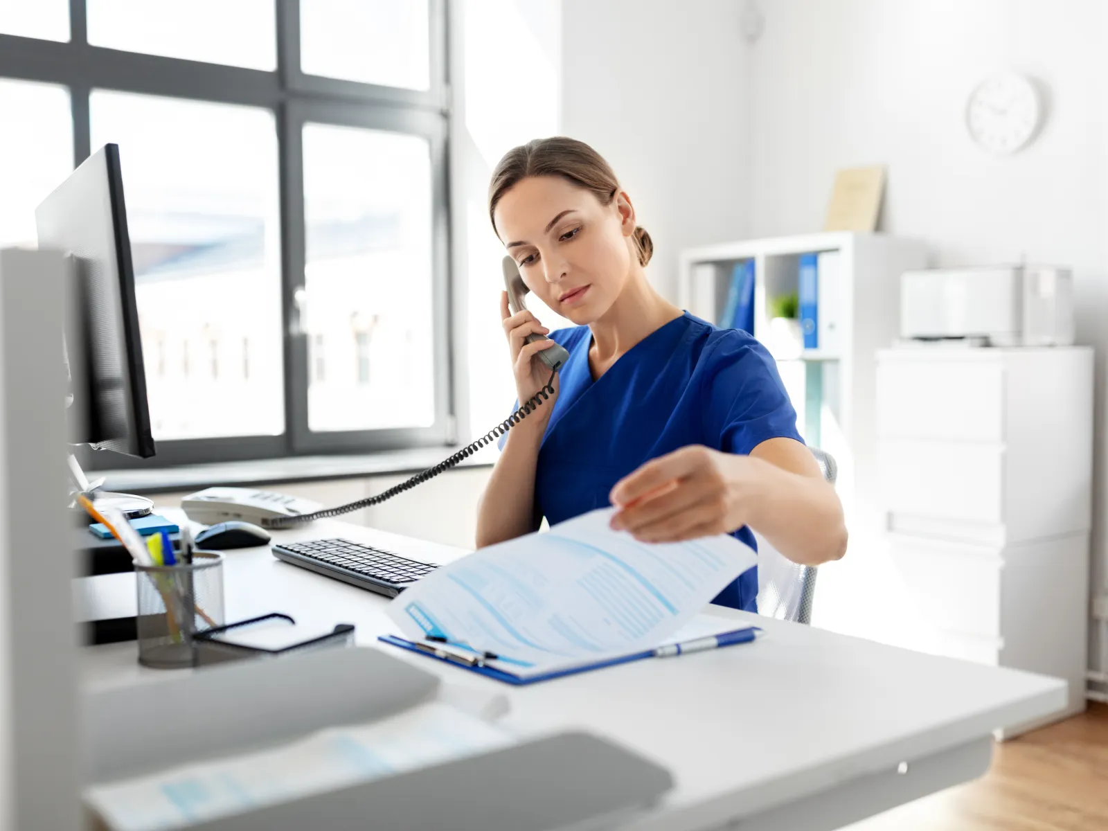 Female healthcare worker in blue scrubs talking on the phone and reviewing documents in a bright office.