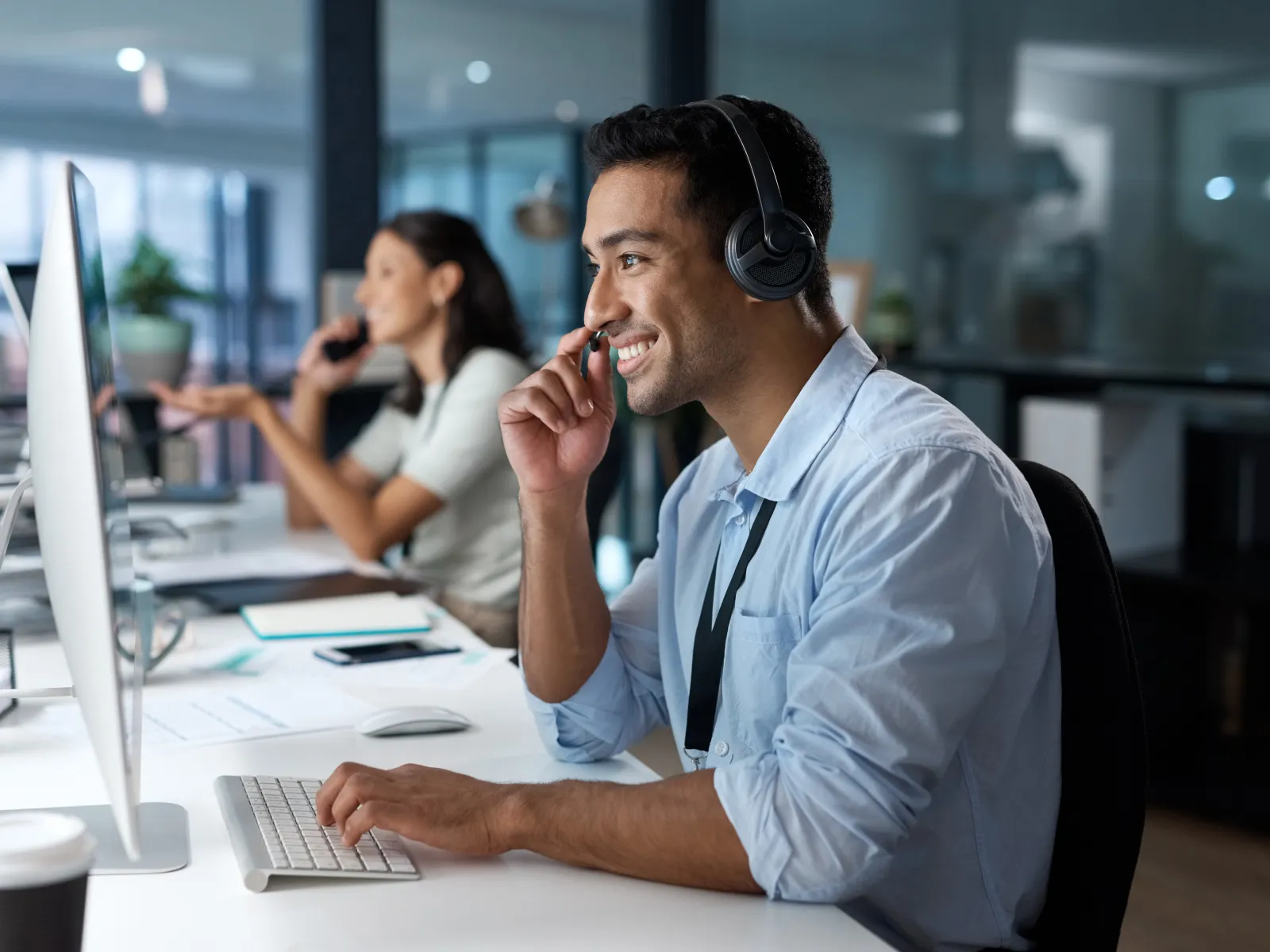 Smiling man wearing headset working at computer in a modern office with colleague in the background.