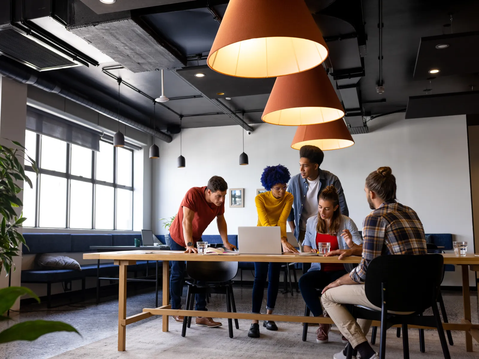 Five diverse coworkers collaborate around a laptop in a modern office with pendant lighting and large windows.