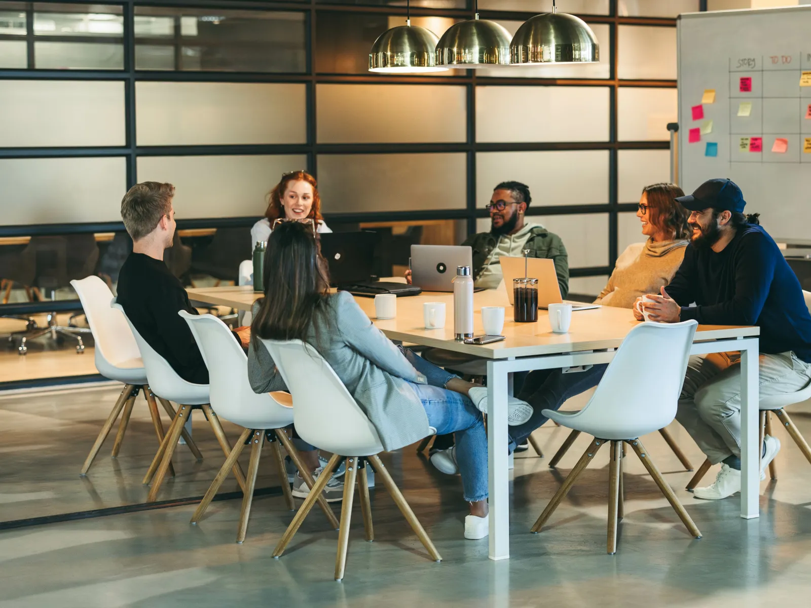 Team meeting in modern office with diverse colleagues discussing projects around a table near a whiteboard with sticky notes.