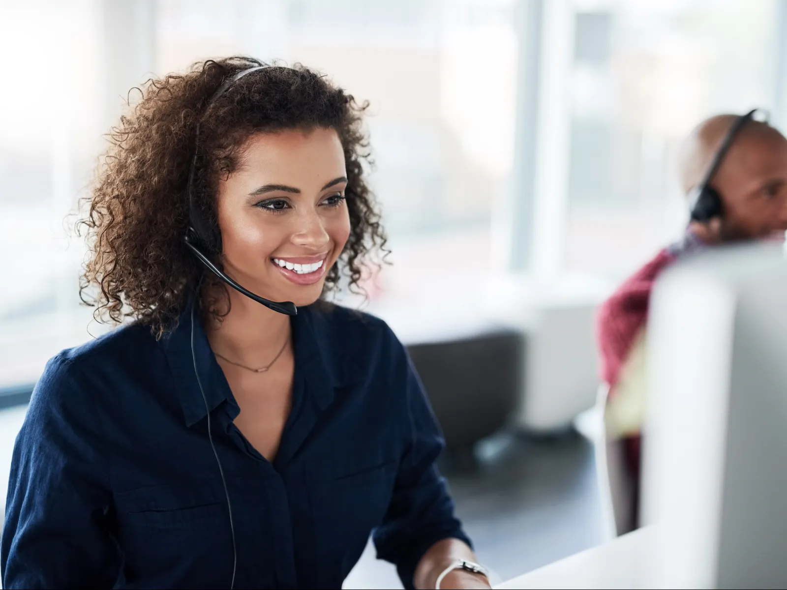 Smiling customer service representative wearing headset working at computer in modern office environment.