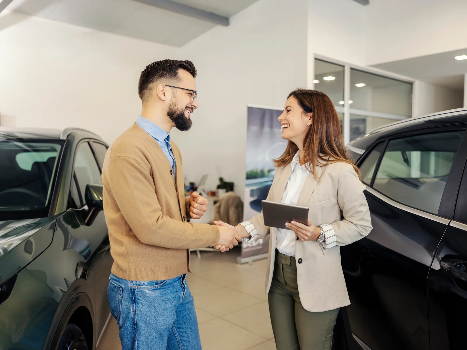Man and woman shaking hands in a car dealership between two black cars, smiling and holding a tablet.