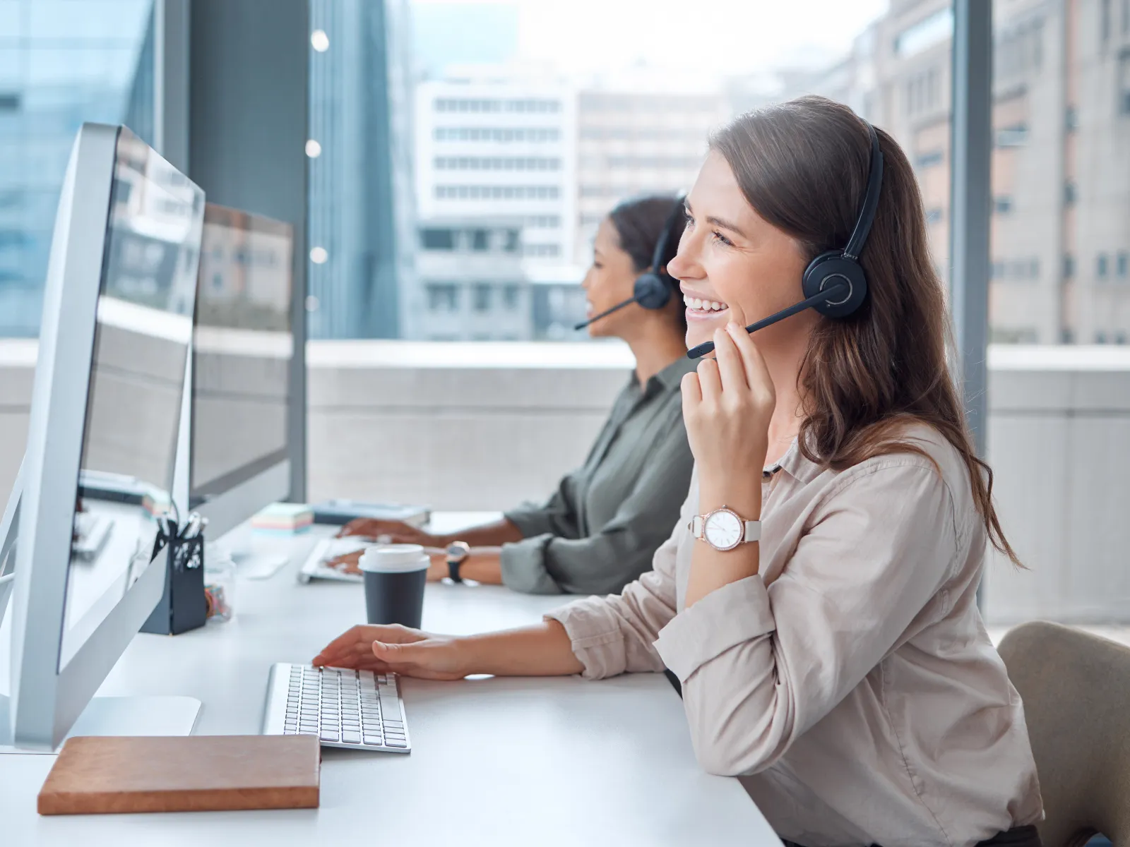 Two customer service representatives wearing headsets working on computers in a modern office with large windows.