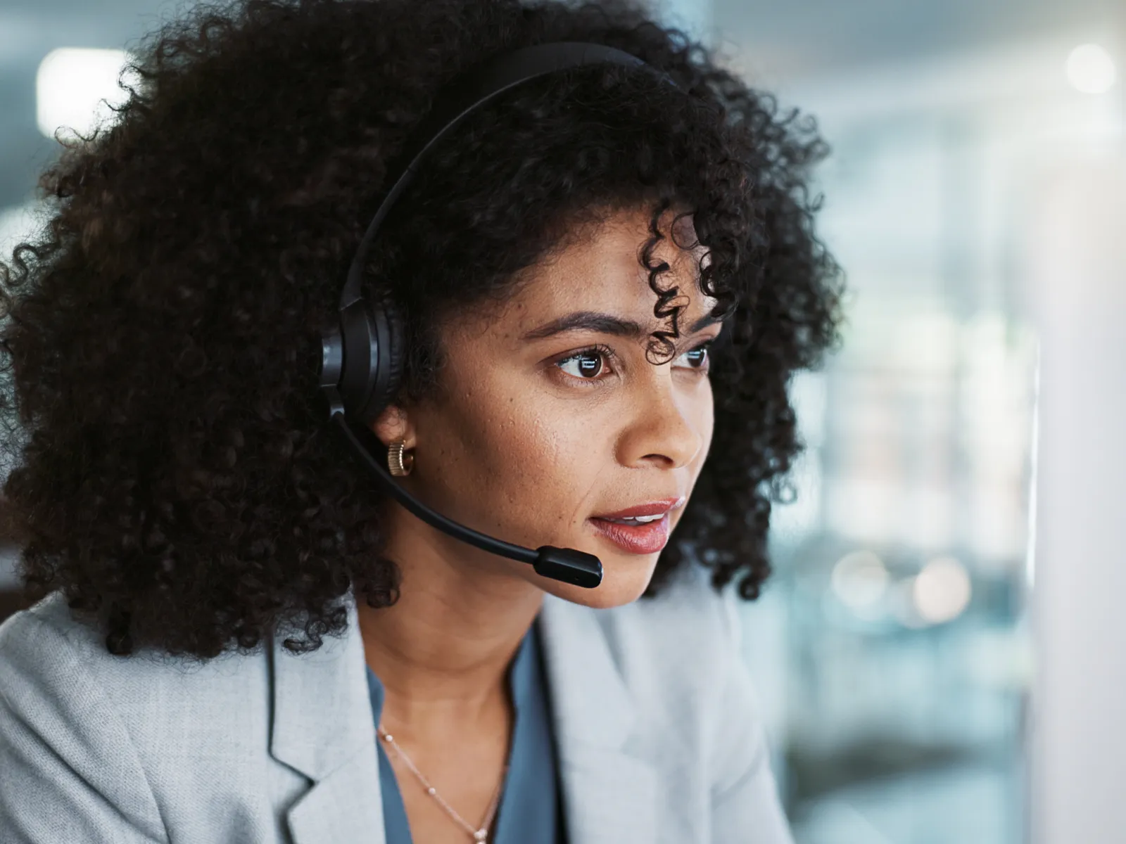Young woman with curly hair wearing a headset and gray blazer working attentively at a computer in an office.
