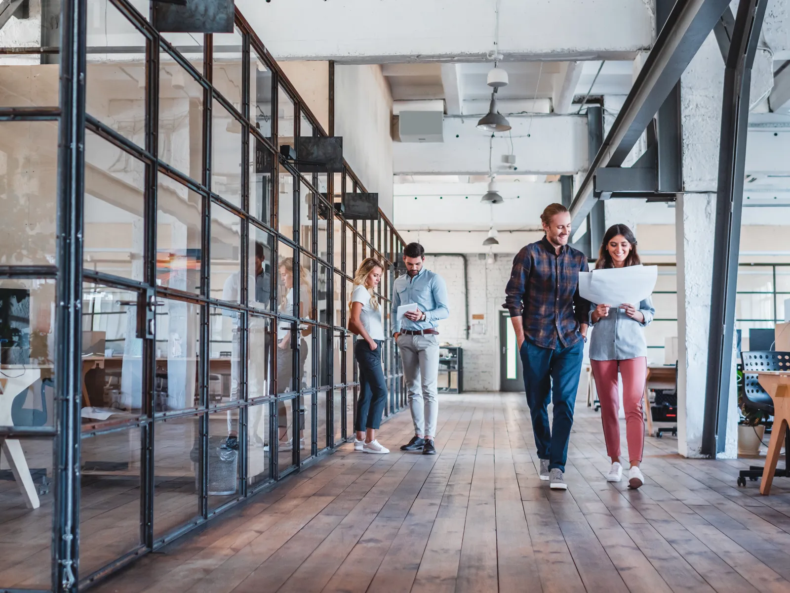 Two colleagues walk and discuss a project in a modern office with glass walls and wooden floors.