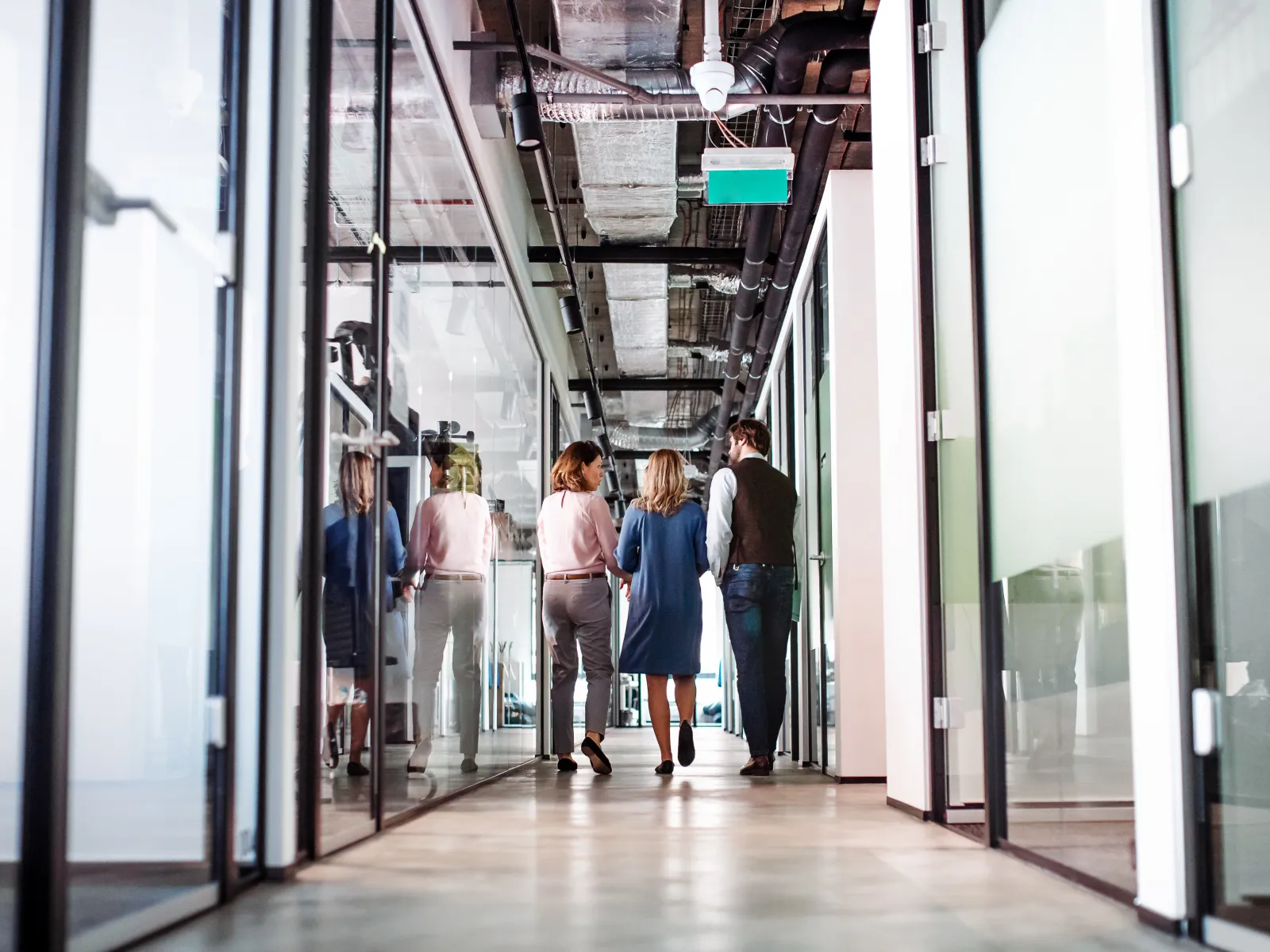 Three professionals walking through a modern glass-walled office corridor with exposed ceiling pipes and lighting.