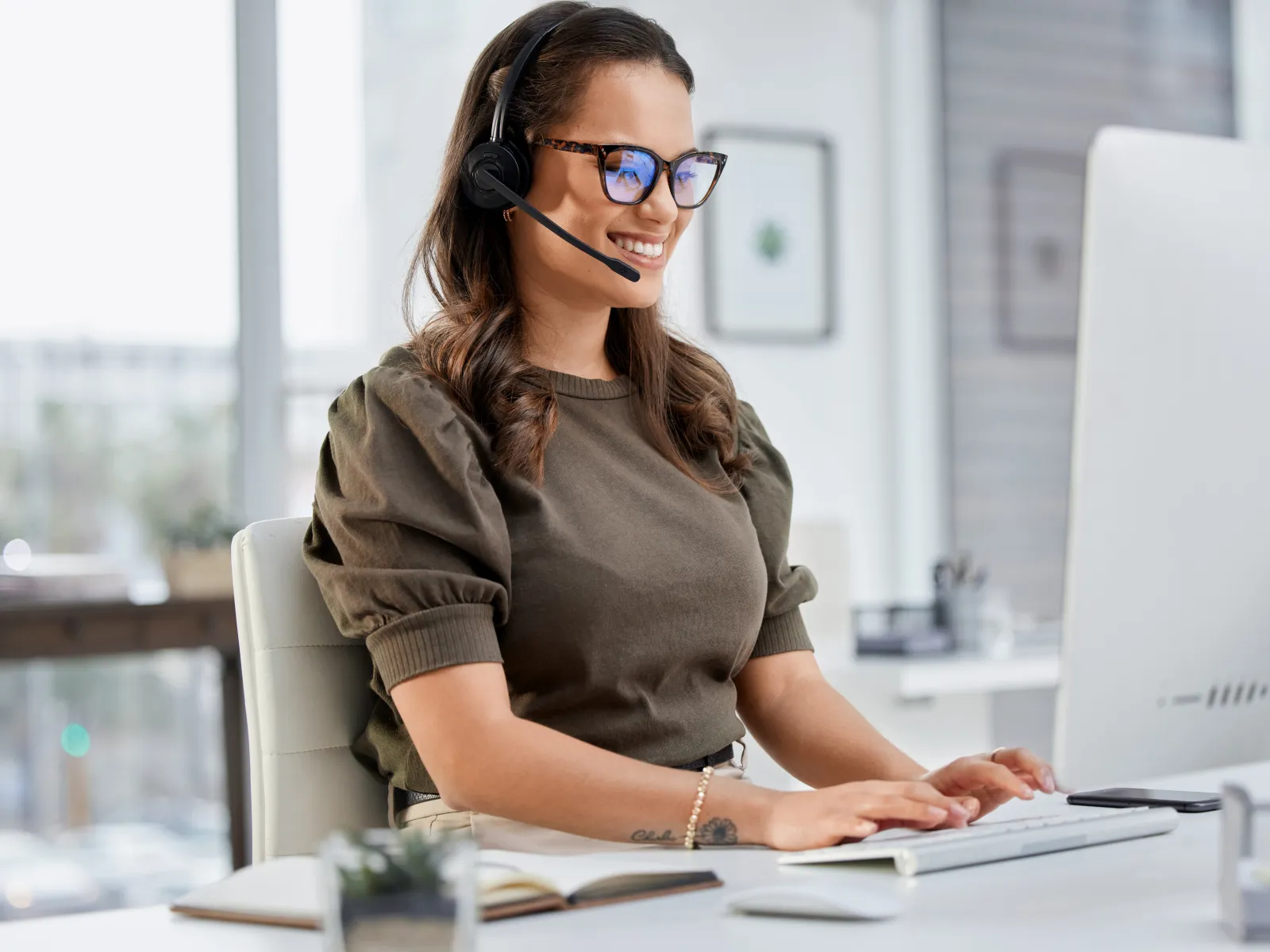 Smiling woman wearing headset working on computer in bright modern office with large window and plants.