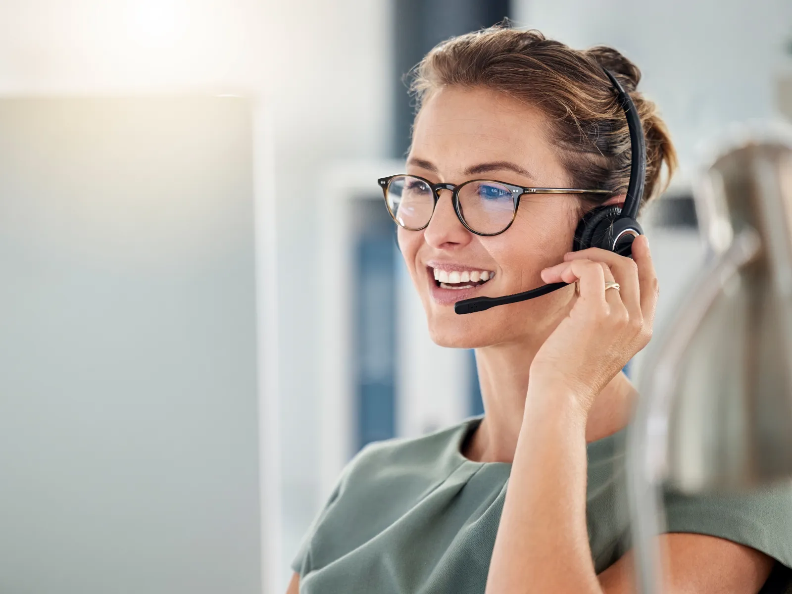 Smiling woman wearing headset and glasses working at computer in modern office environment.