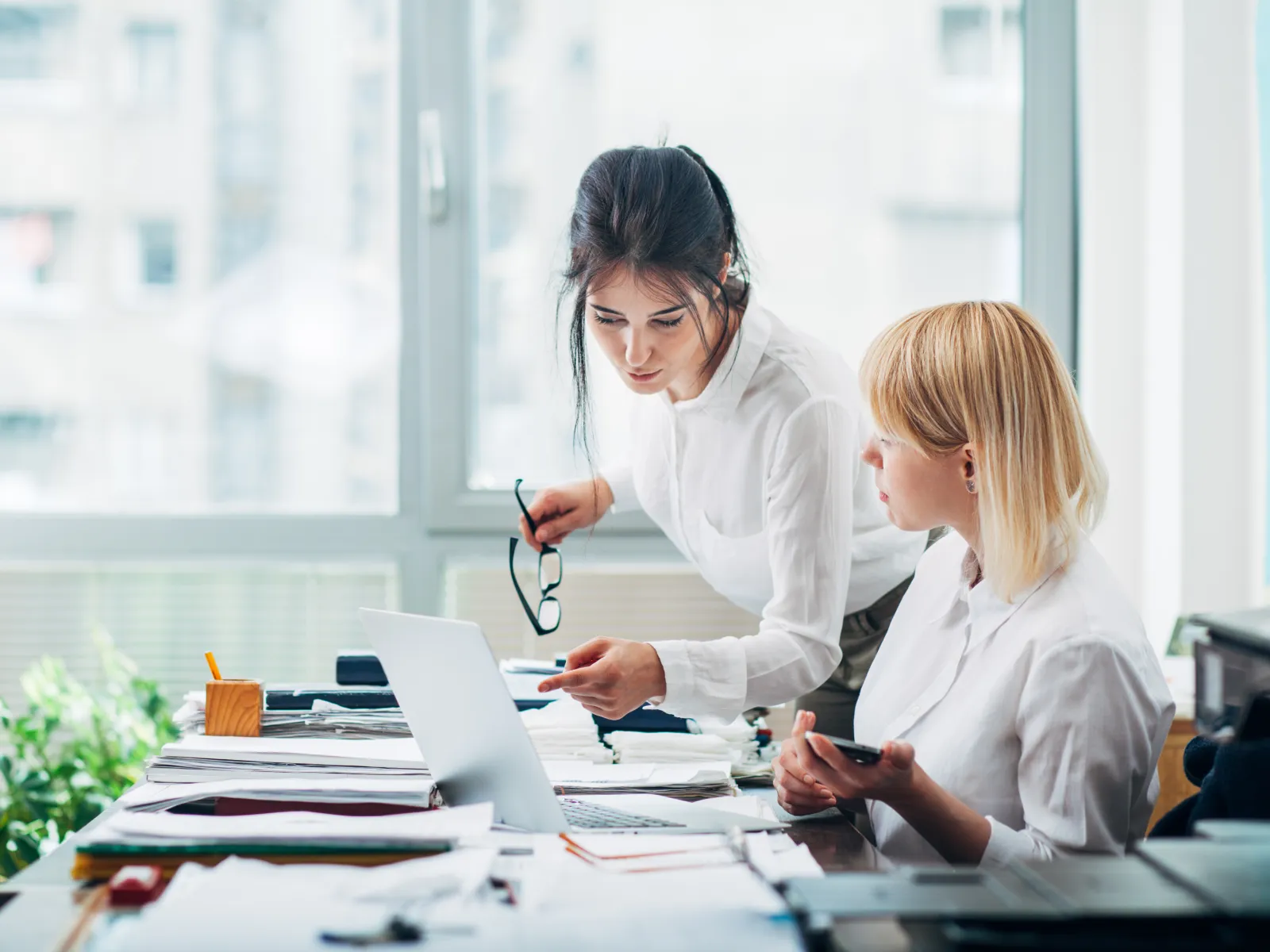 Two women collaborating on a project, discussing notes and laptop screen in a bright office workspace.
