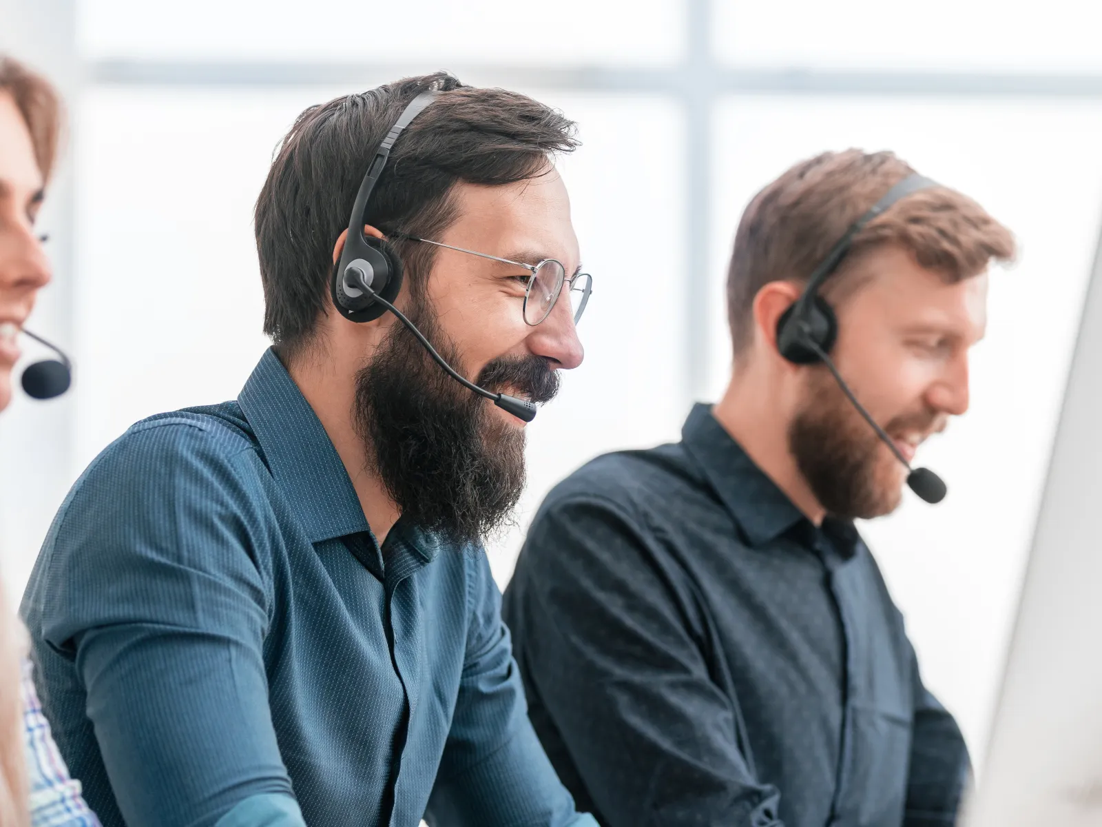 Two male customer service representatives with headsets smiling and working at computers in a bright office.