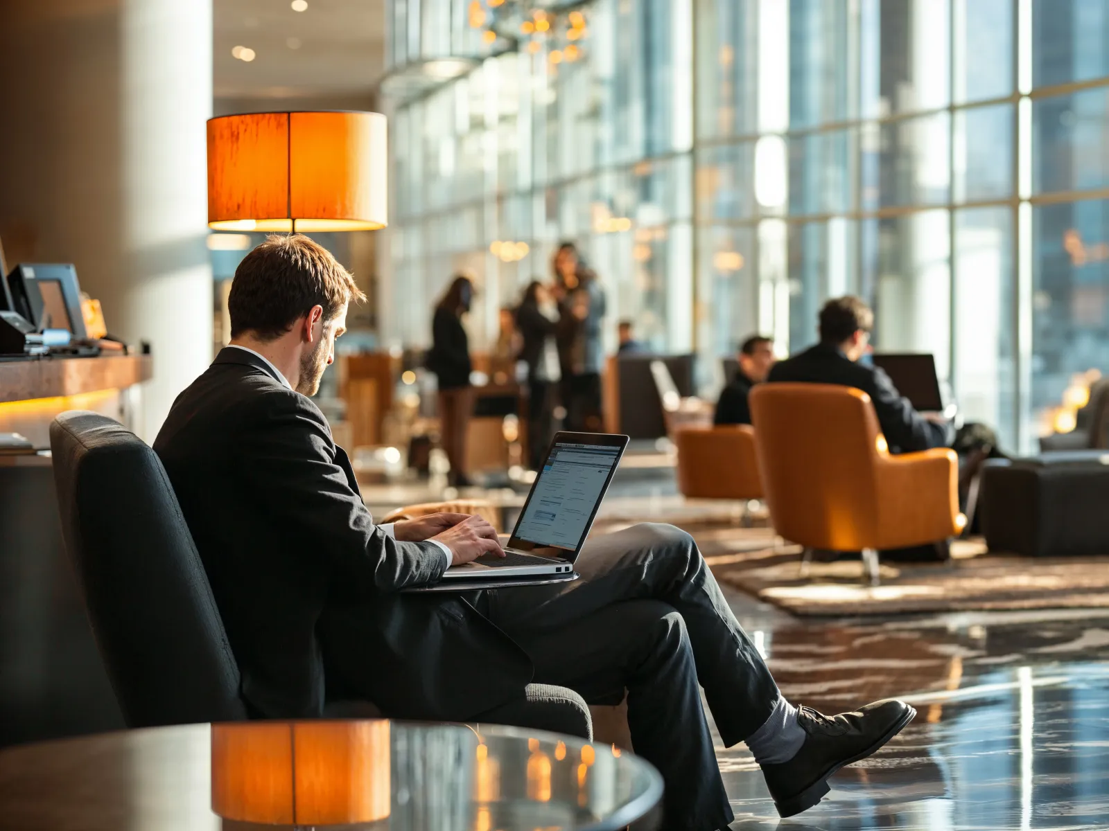 Businessman working on laptop in modern office lounge with large windows and warm lighting.