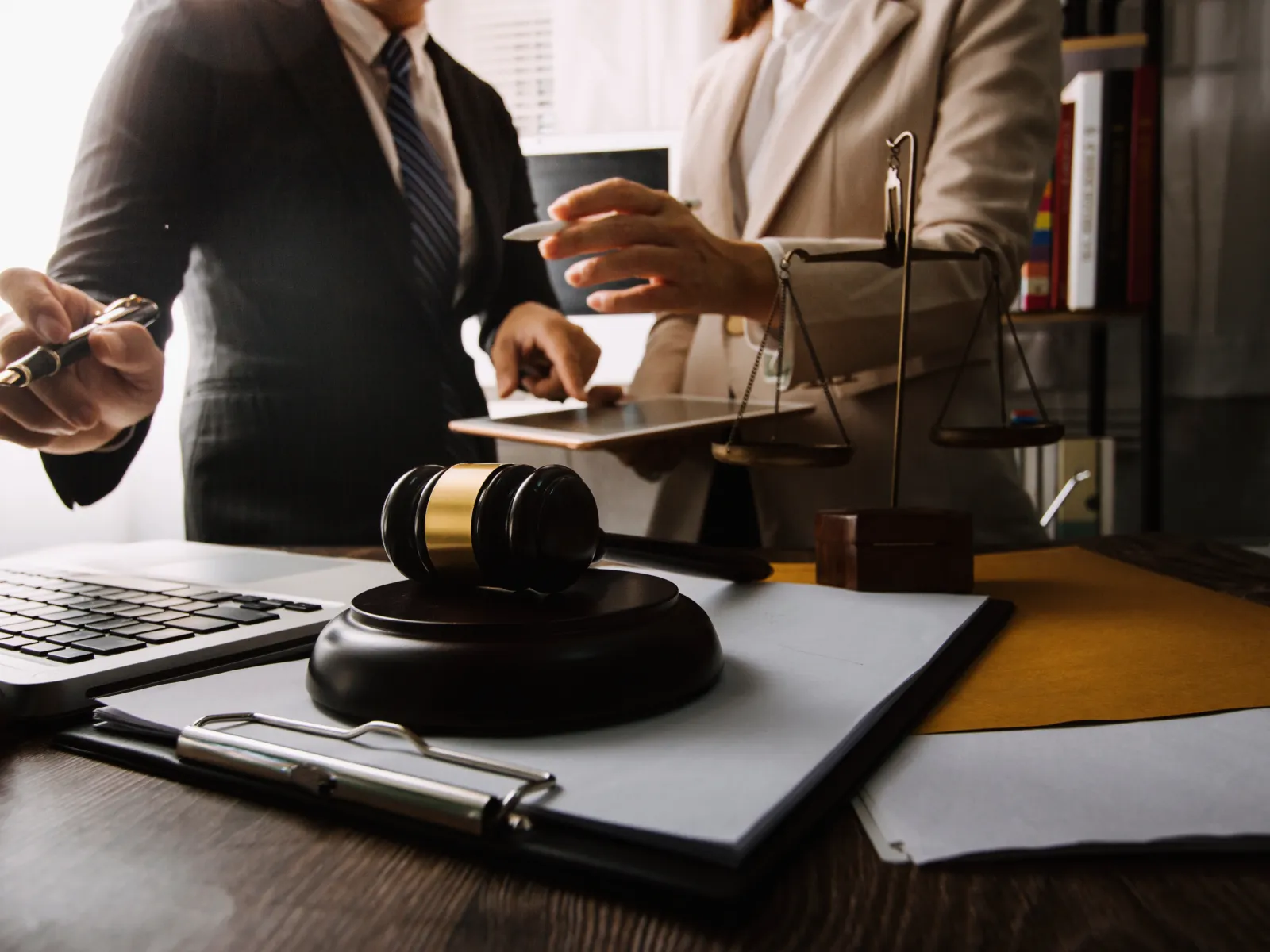 Legal professionals discussing case files beside a gavel and scales of justice on a desk with laptop and documents.