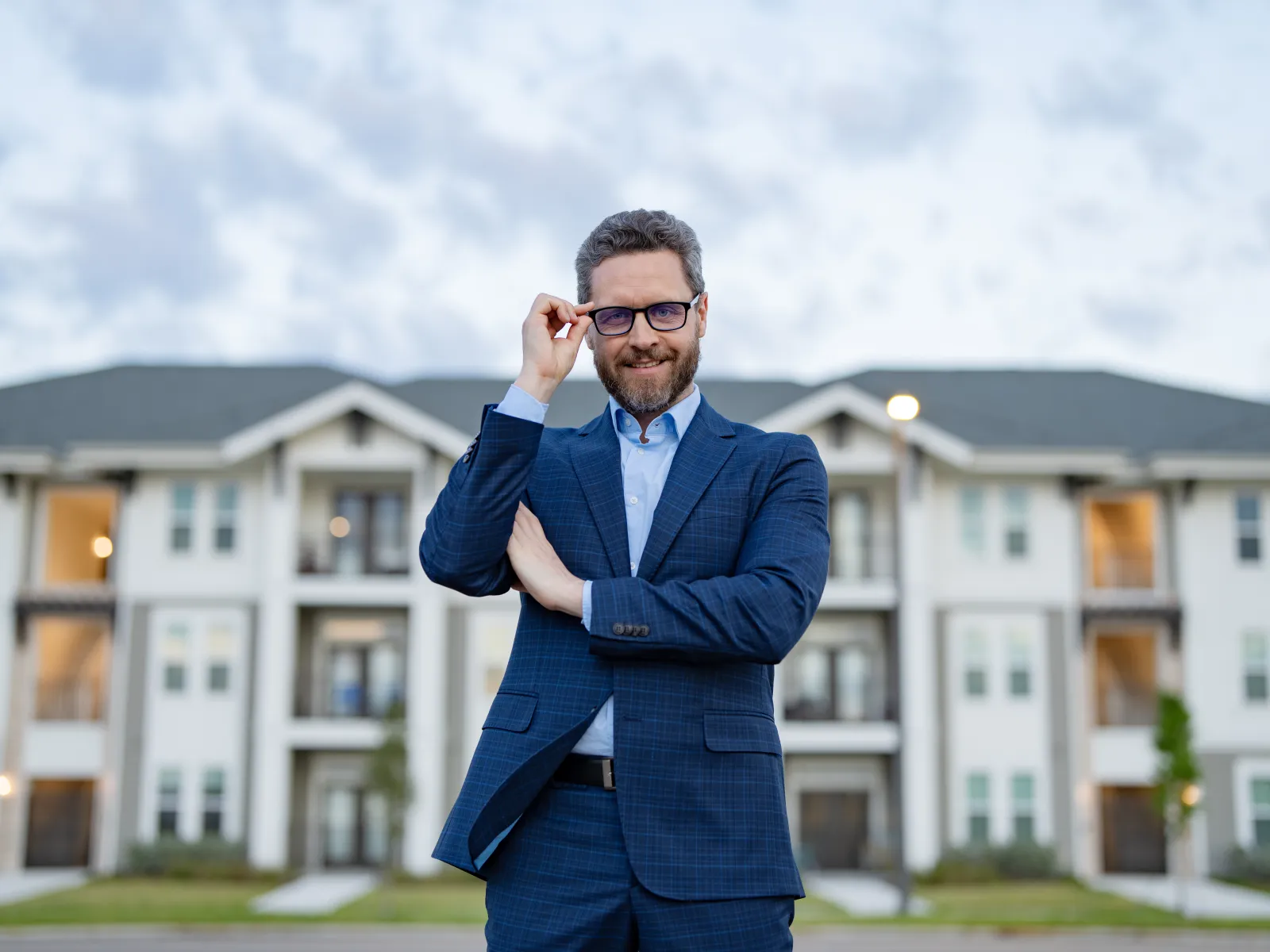 Confident man in suit adjusting glasses standing in front of a modern apartment building at dusk.
