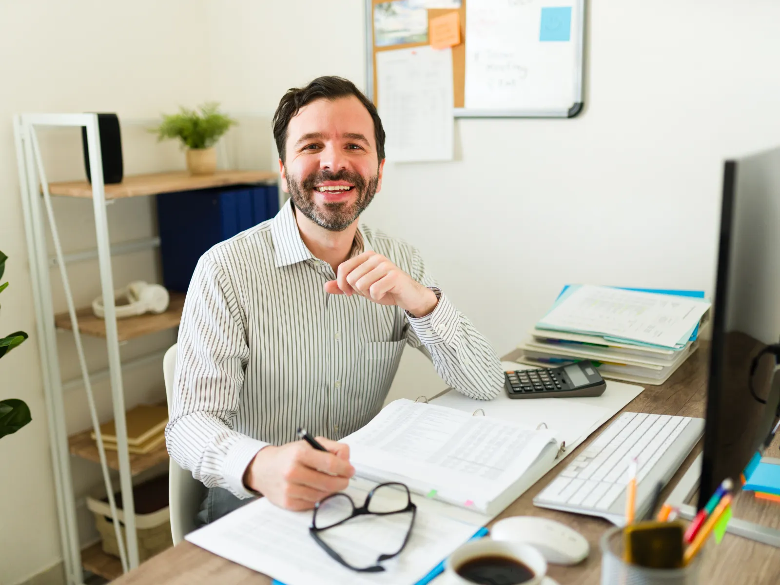 Smiling man working at desk with documents, computer, calculator, and glasses in bright office space