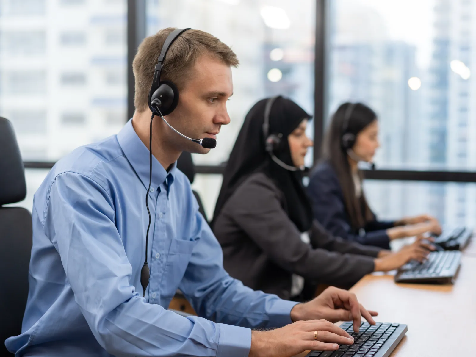 Diverse call center agents wearing headsets working on computers in a modern office with city view.