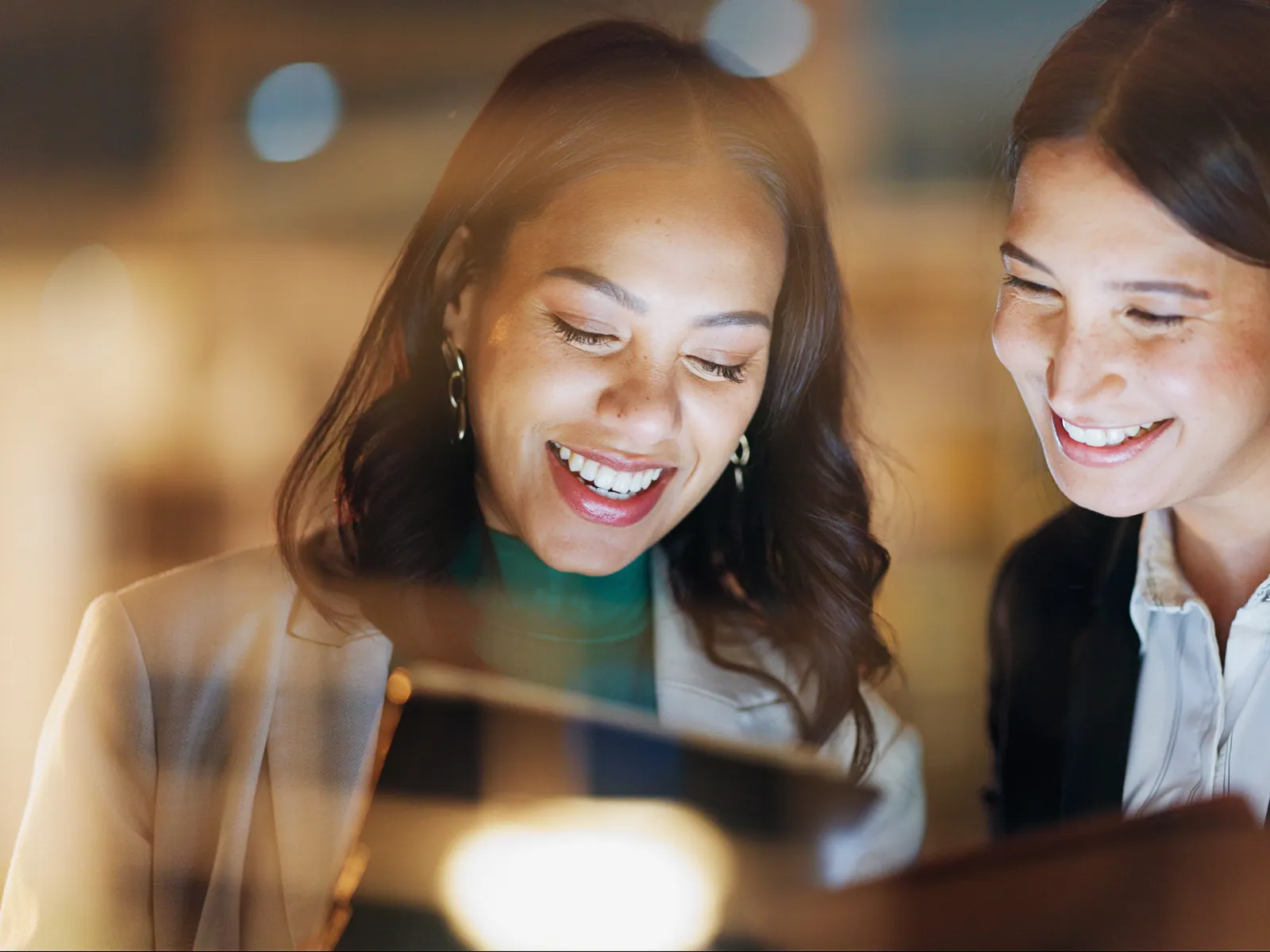 Two professional women smiling and looking at a document in a warm office setting with soft lighting.