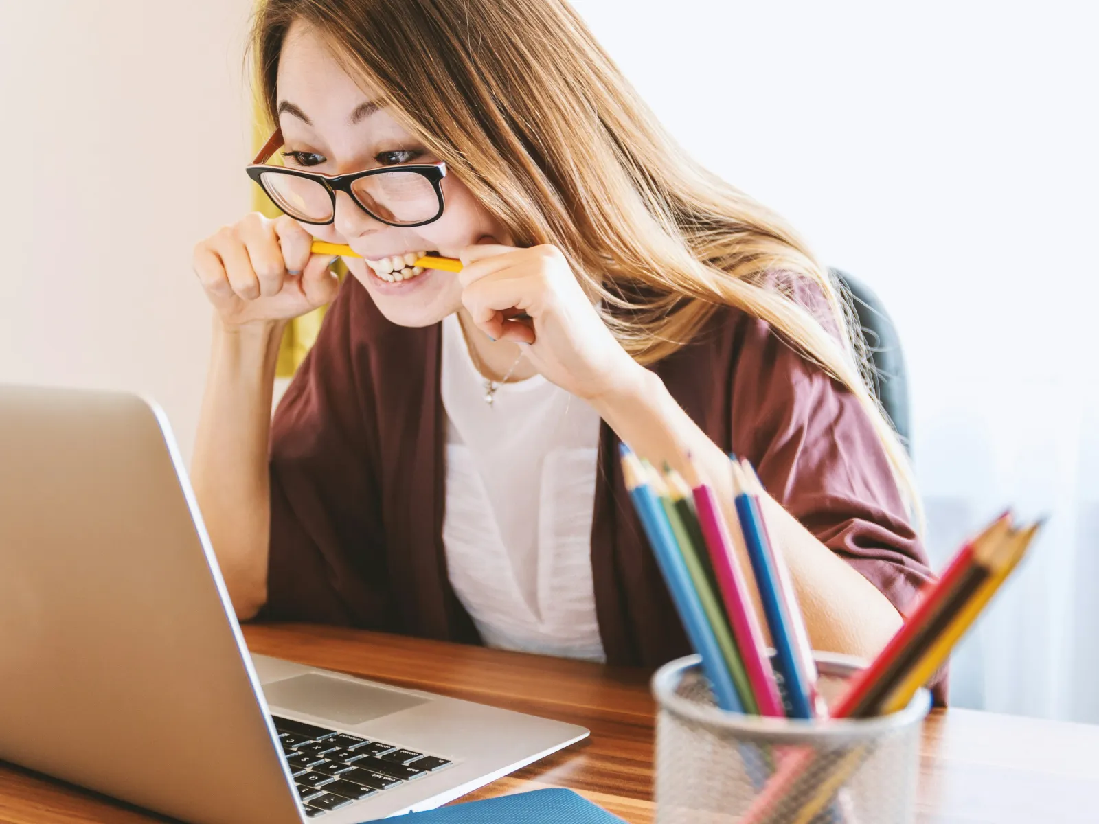 Stressed young woman biting pencil while working on laptop at wooden desk with colorful pencils in holder.