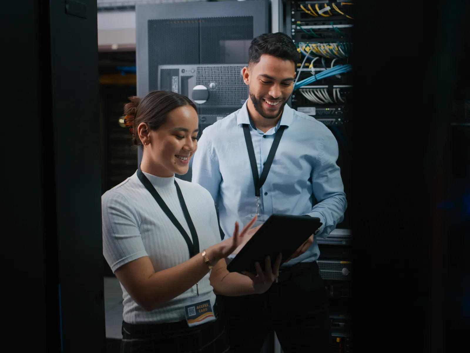 Two IT professionals smiling and reviewing data on a tablet inside a server room with network cabinets.