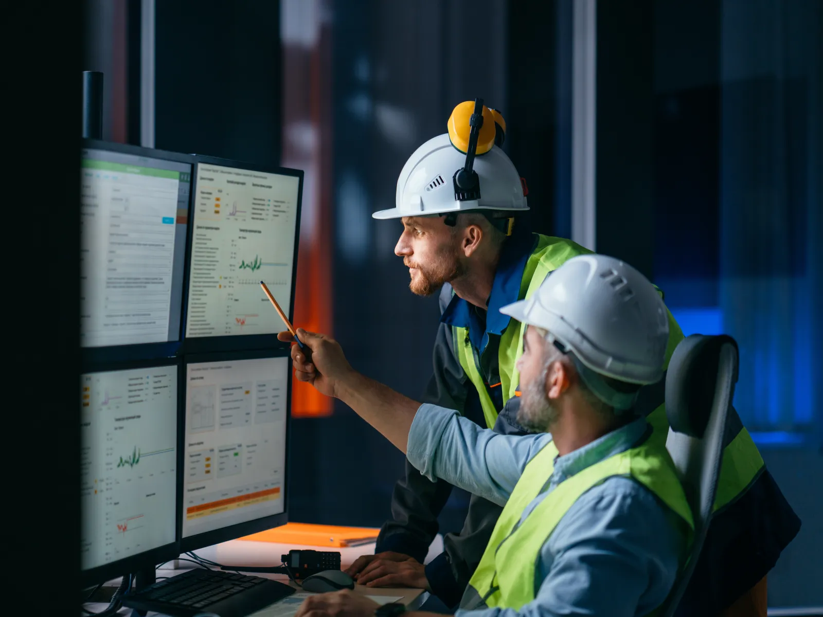 Engineers in hard hats analyzing data on multiple monitors in a dimly lit control room.