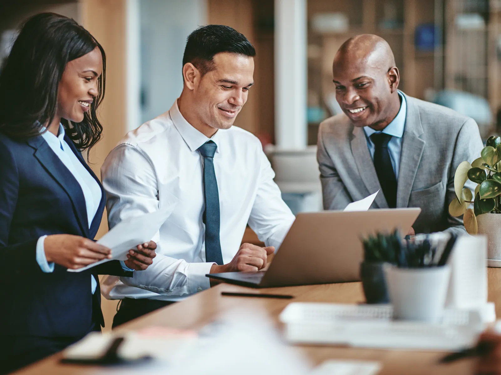 Three diverse business professionals smiling while collaborating on a project using a laptop and documents in an office.