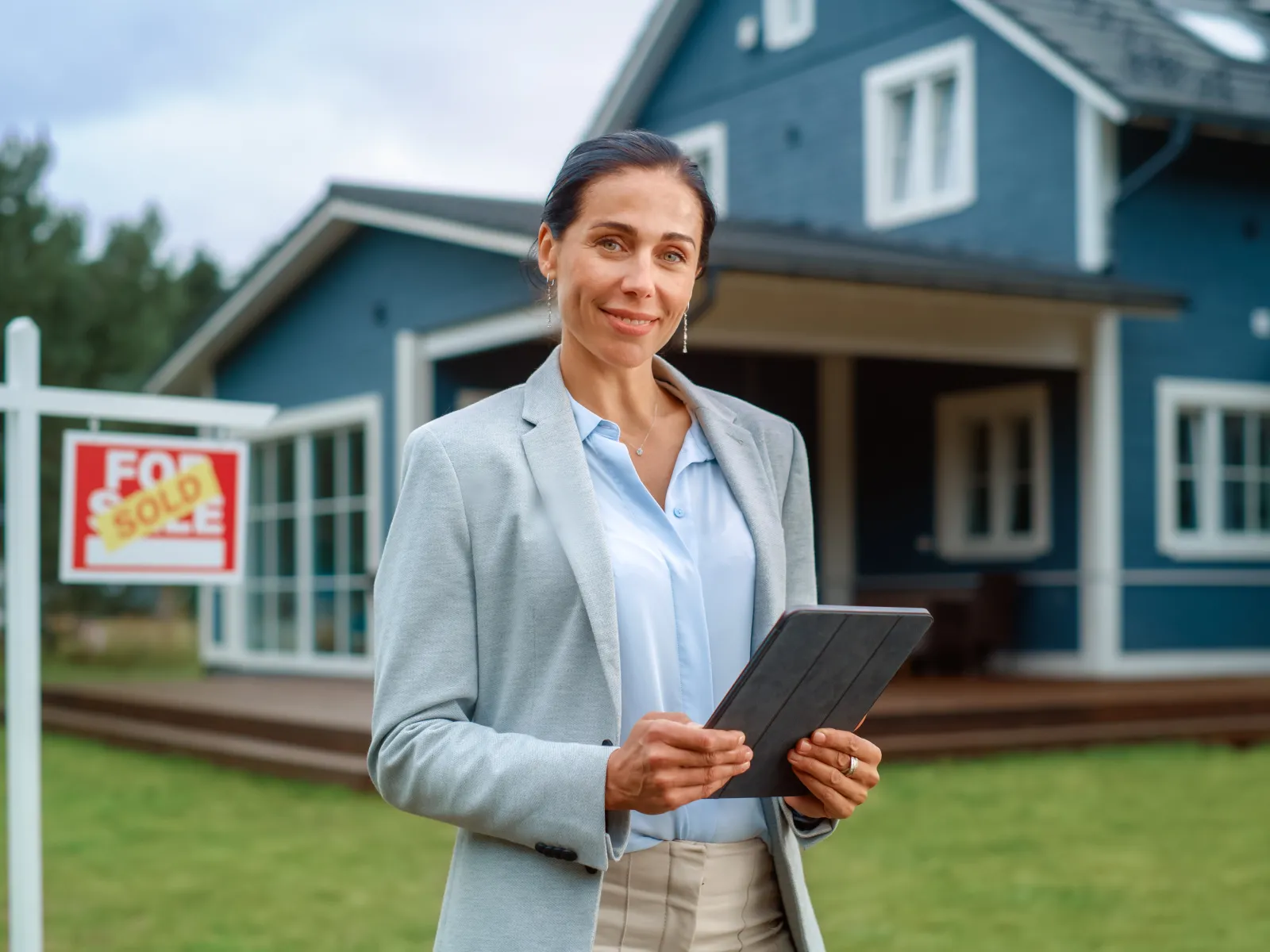 Confident female real estate agent holding tablet in front of sold house with a sold sign on lawn