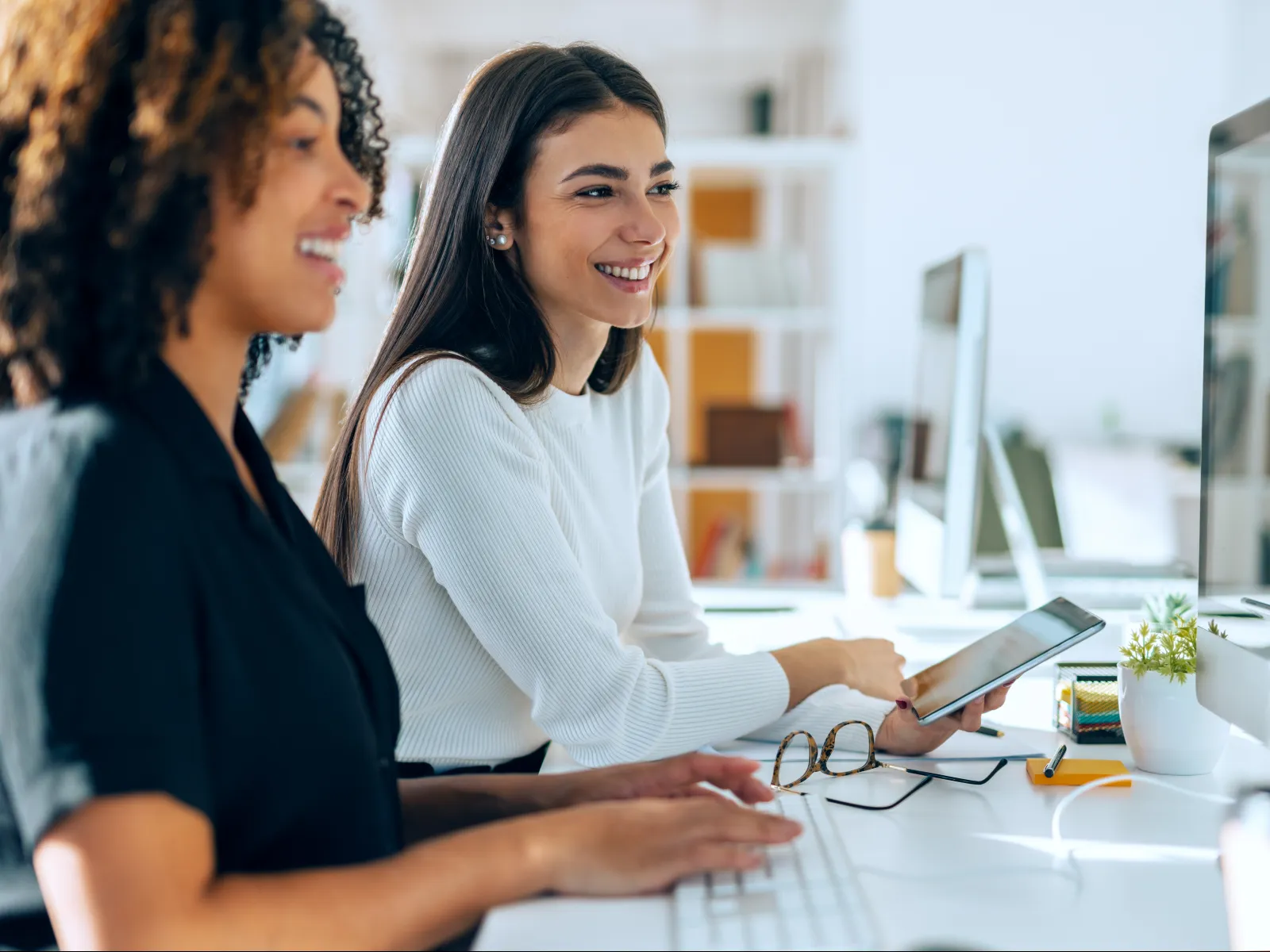 Two colleagues collaborating happily at desk with computer and tablet in modern office setting