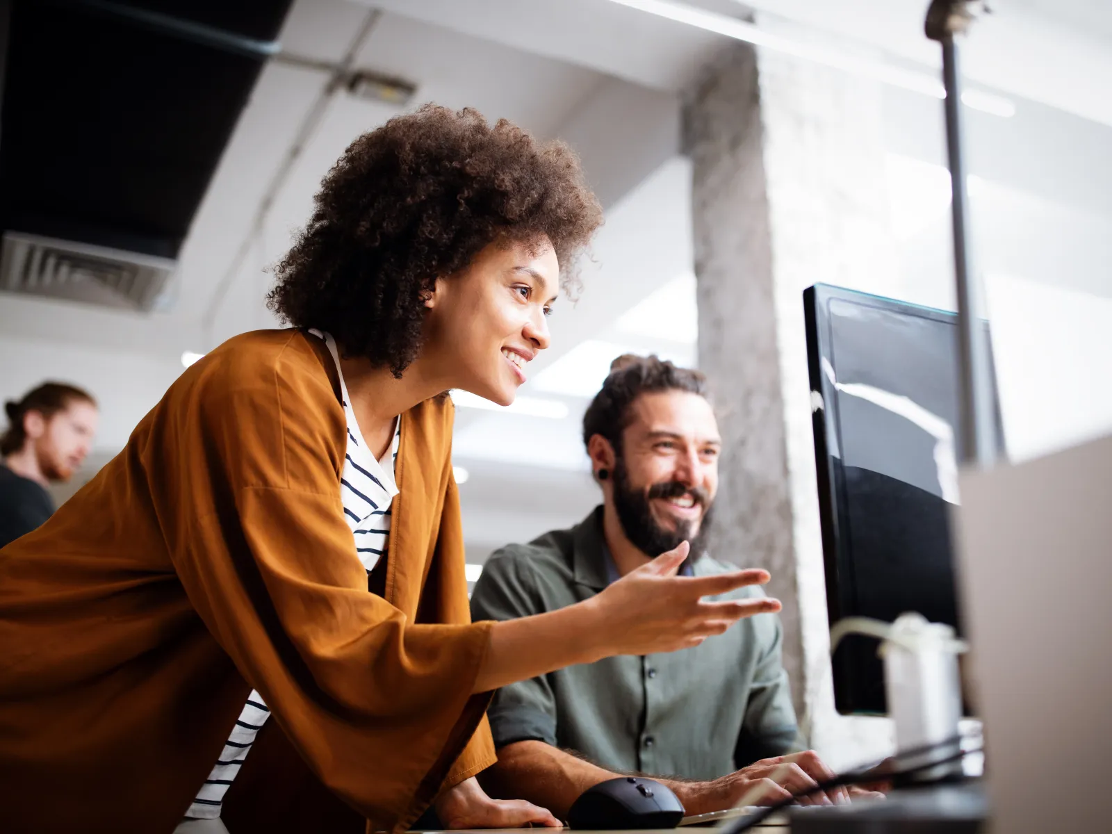 Two coworkers smiling and collaborating while looking at a desktop computer screen in a modern office.