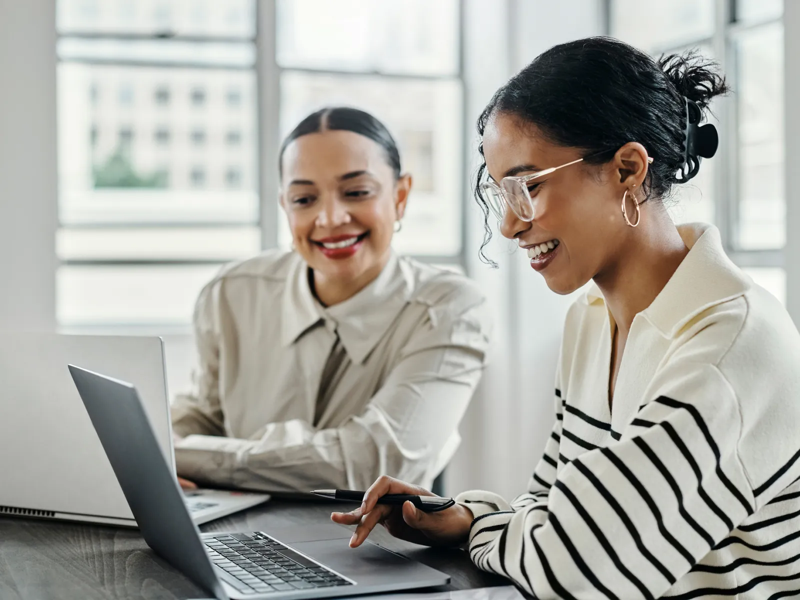 Two women collaborating on laptops in a bright modern office, smiling and discussing work together.