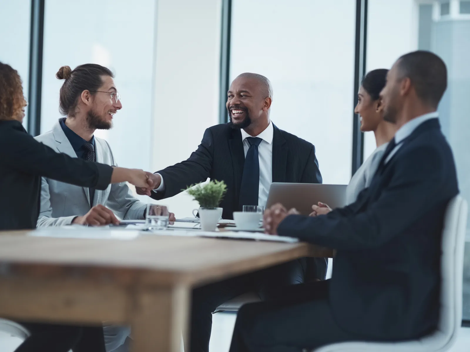 Business professionals shaking hands during a meeting in a modern office setting with laptops and documents.
