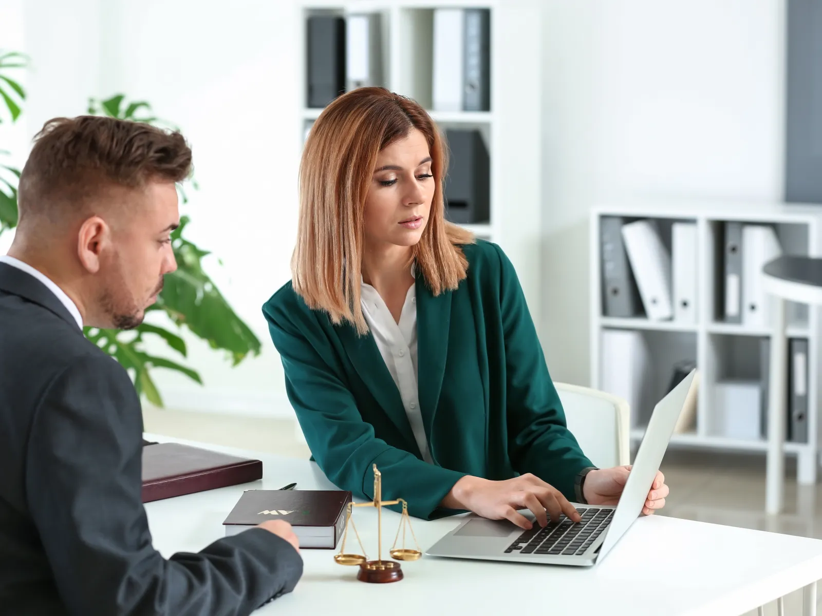Businesswoman explains legal information to businessman during meeting at office with laptop and scales of justice.