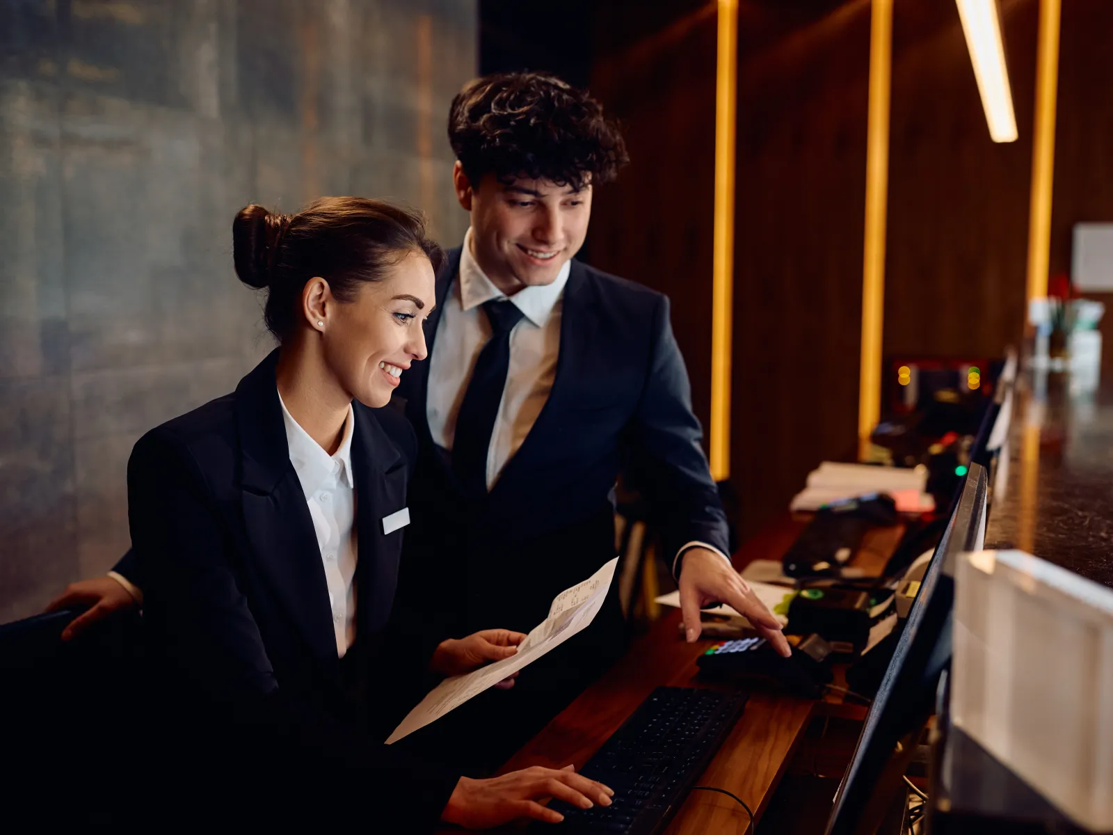 Two hotel receptionists in suits assisting guests at the front desk with a computer and documents.