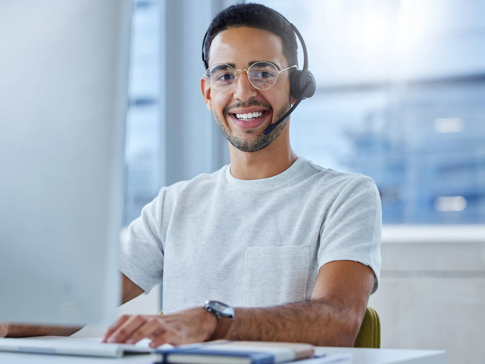 Smiling customer service agent wearing headset and glasses working at computer in modern office.