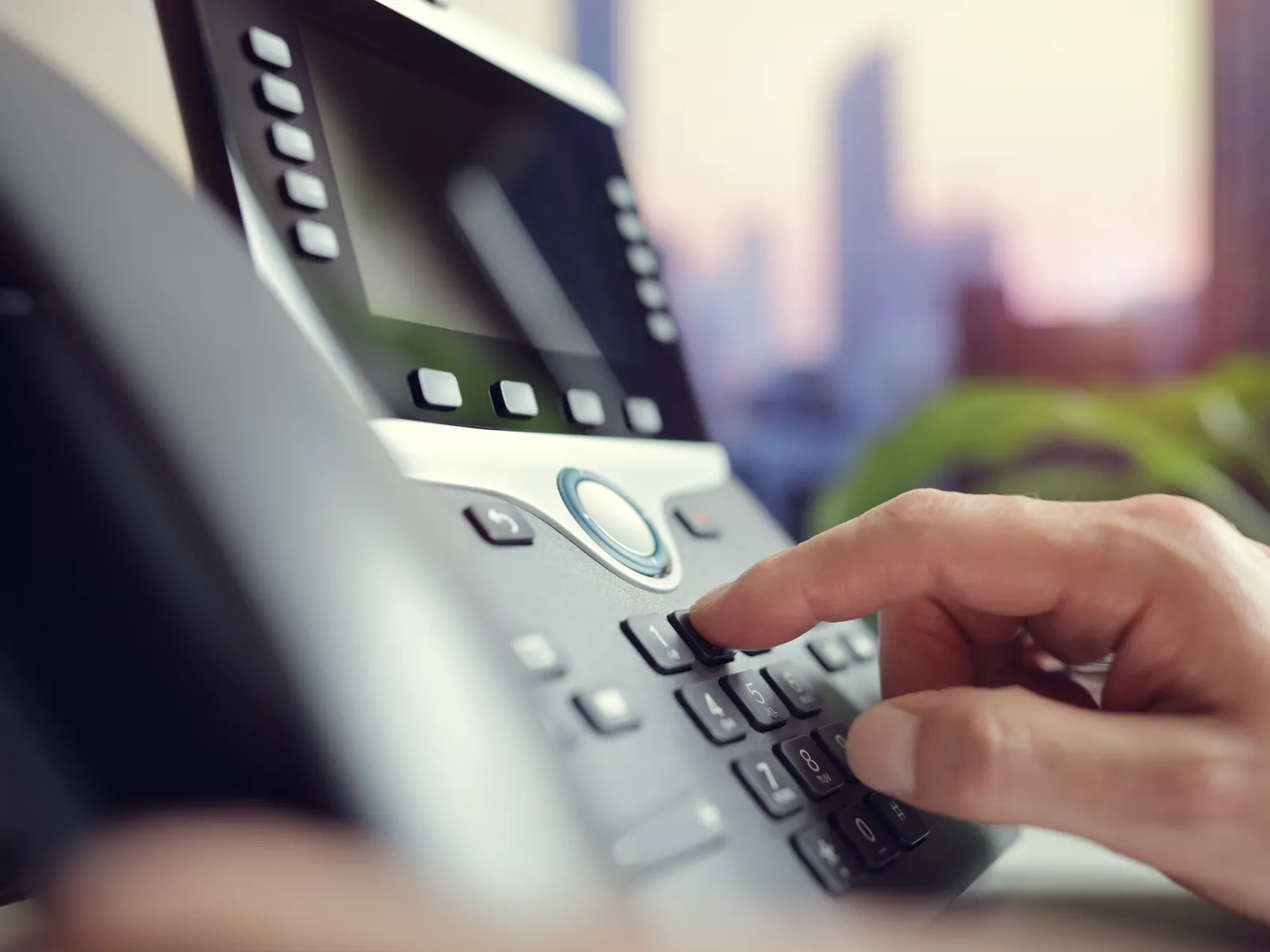 Close-up of a hand dialing a number on a modern office telephone with a blurred city background.