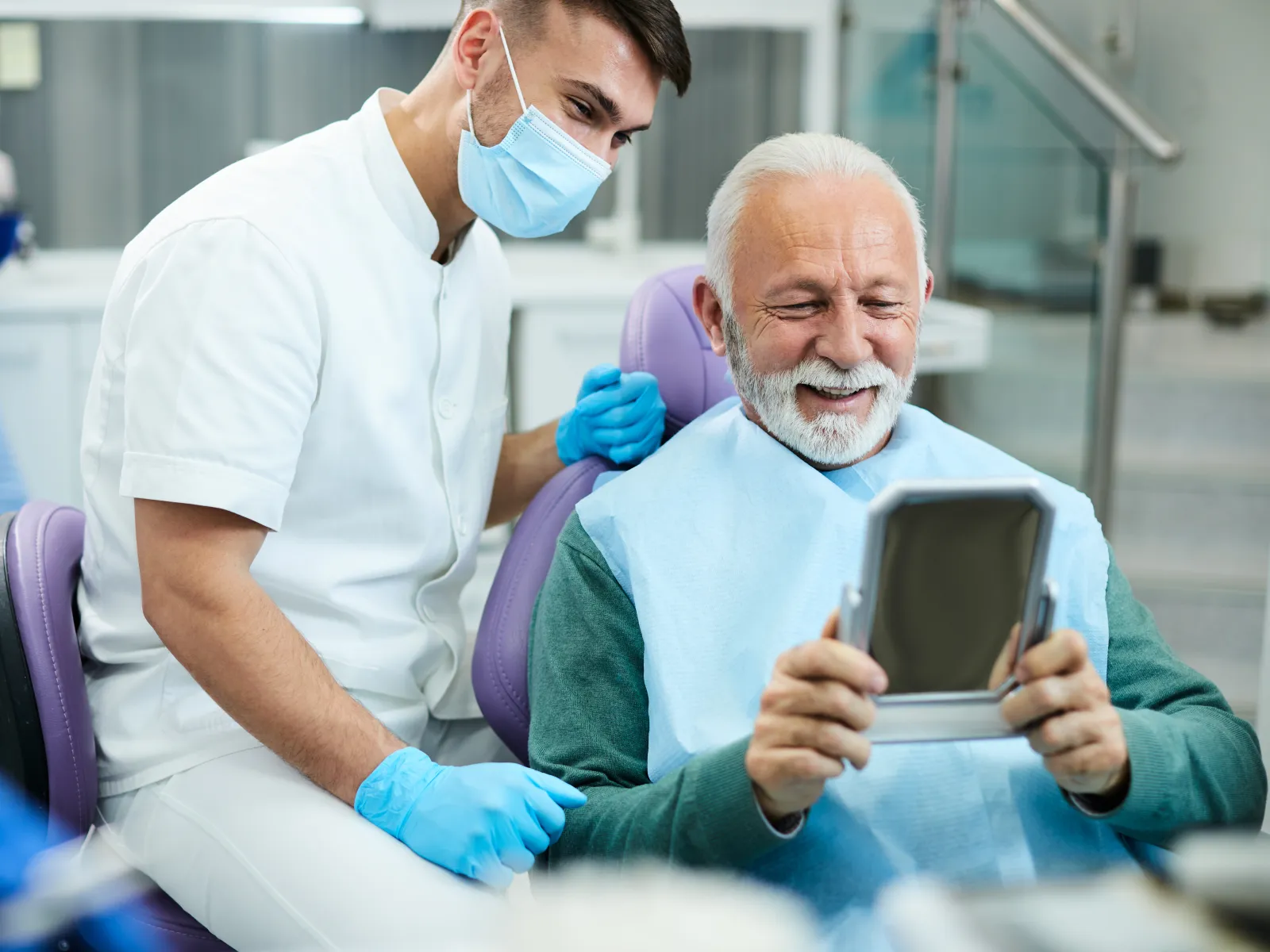 Dentist in mask with gloves talks to smiling senior man holding mirror in dental clinic