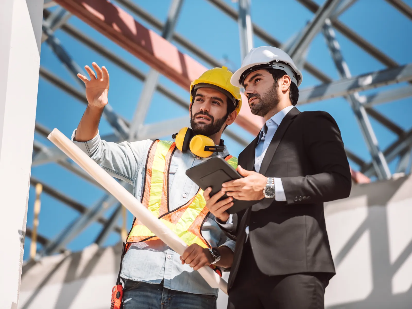 Construction worker and engineer discussing plans at building site with metal framework under blue sky
