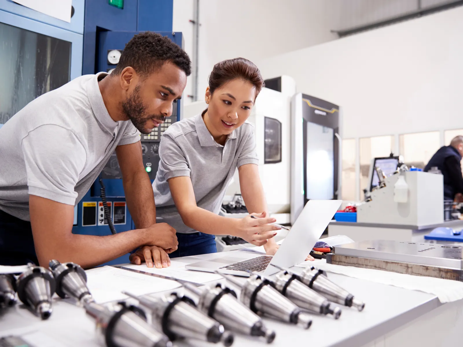 Two engineers review plans on a laptop in a manufacturing workshop with metal parts and machinery.