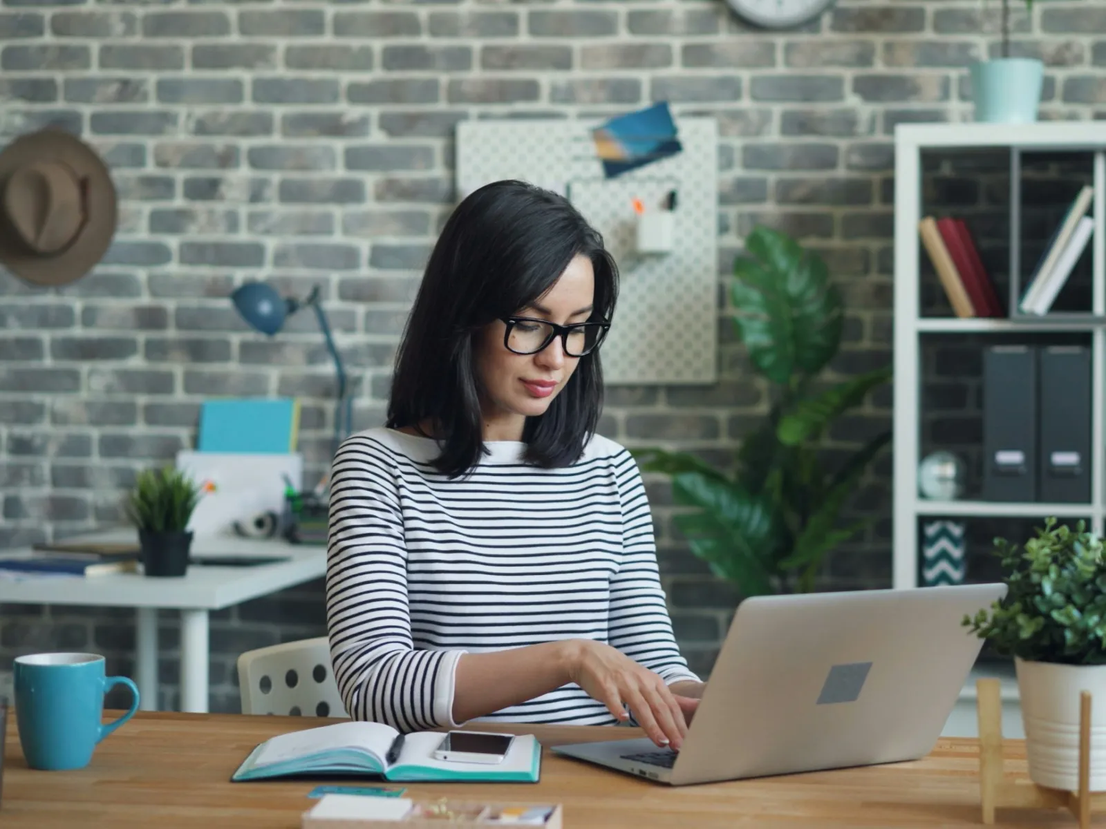 Young woman with glasses working on a laptop at a wooden desk with books and plants in a modern office.