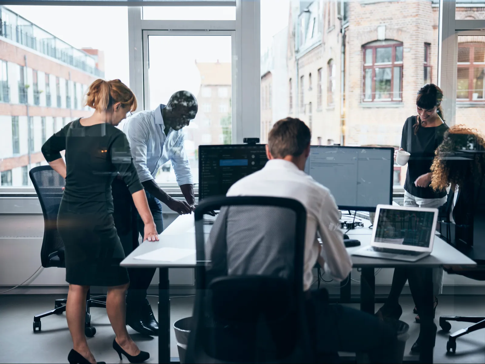 A diverse team collaborates around a modern office table with multiple computers and large windows.