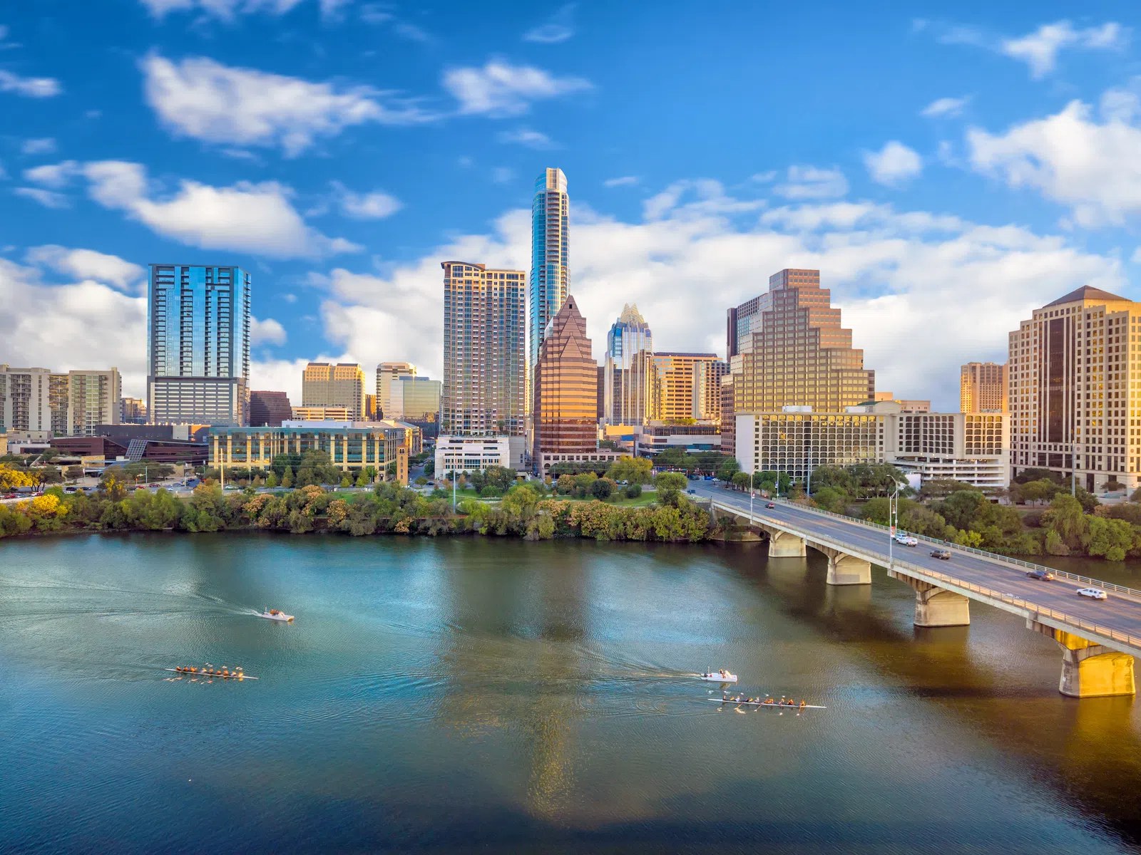 Austin city skyline with high-rise buildings, a bridge over the river, and boats rowing on water under a blue sky.