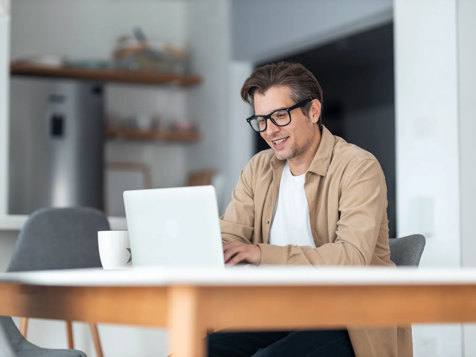 Man in glasses working on a laptop at a modern kitchen table with a cup of coffee nearby.