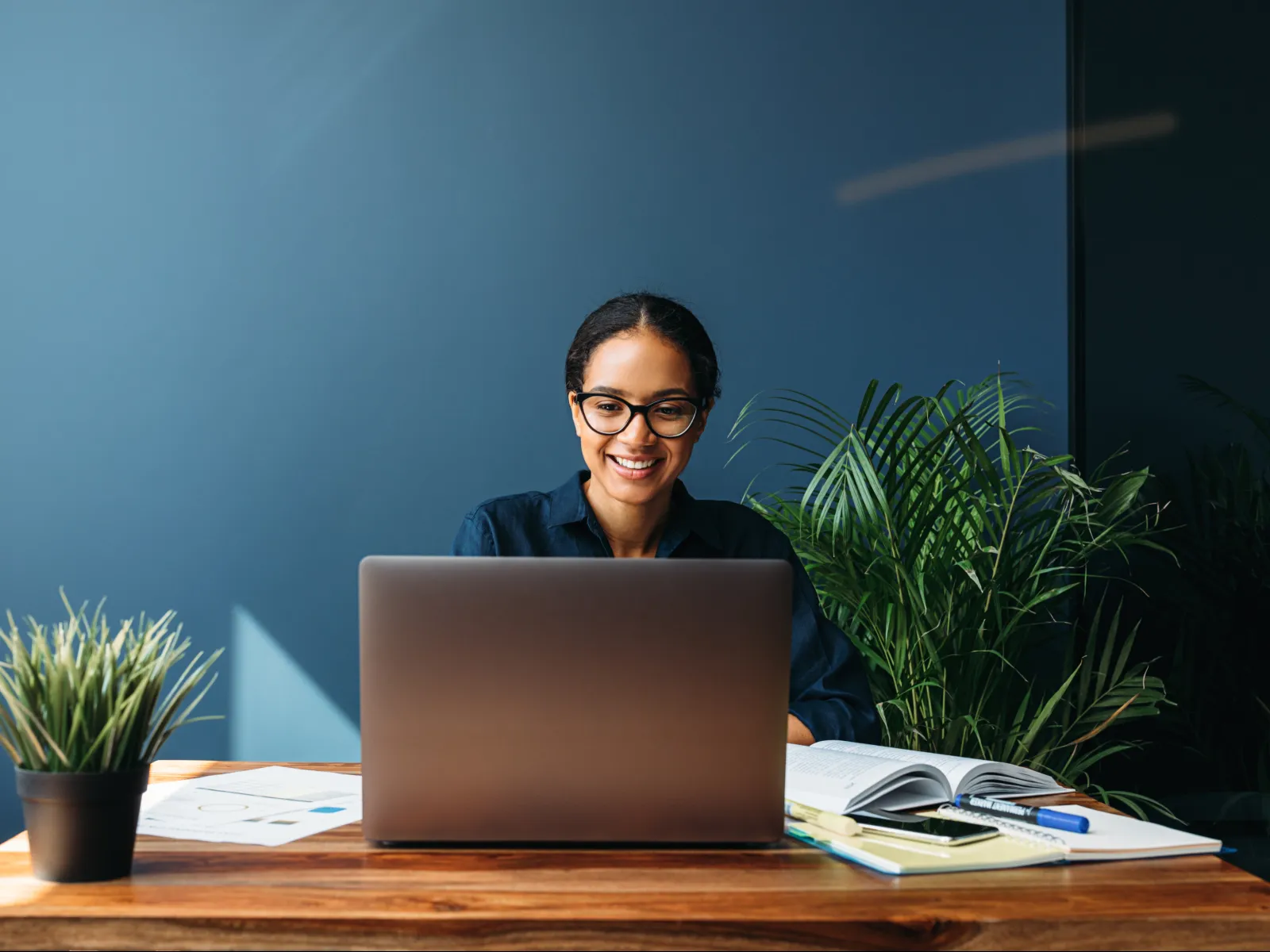 Smiling woman with glasses working on a laptop at a wooden desk with plants and stationery.