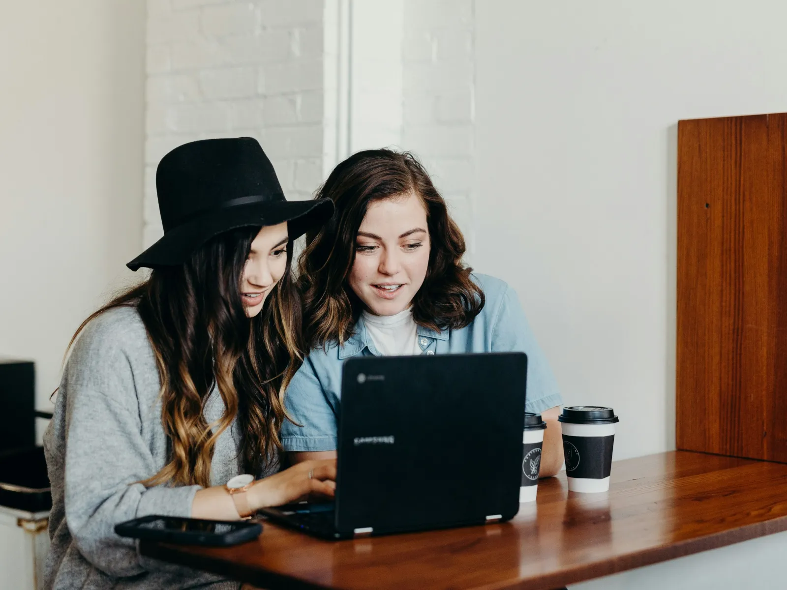 Two women collaborating on a laptop at a cozy cafe with a coffee cup nearby.