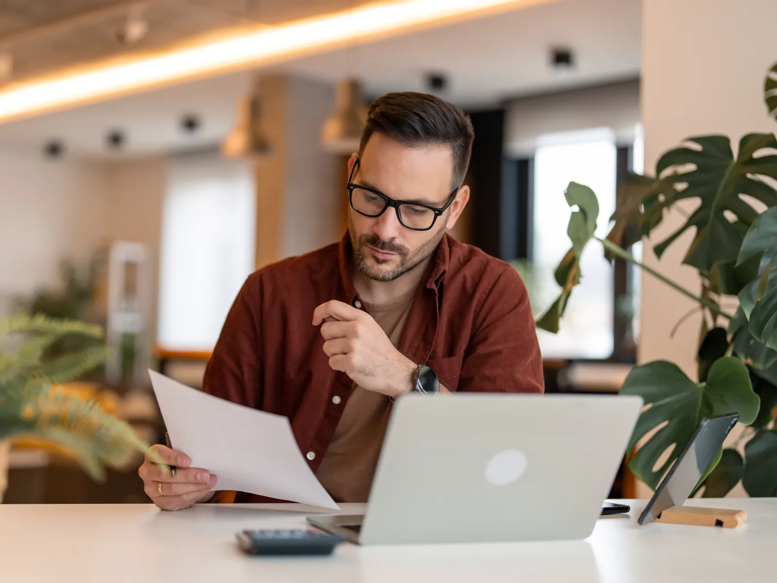 Man in glasses reviewing documents while working on a laptop in a modern office setting with plants.