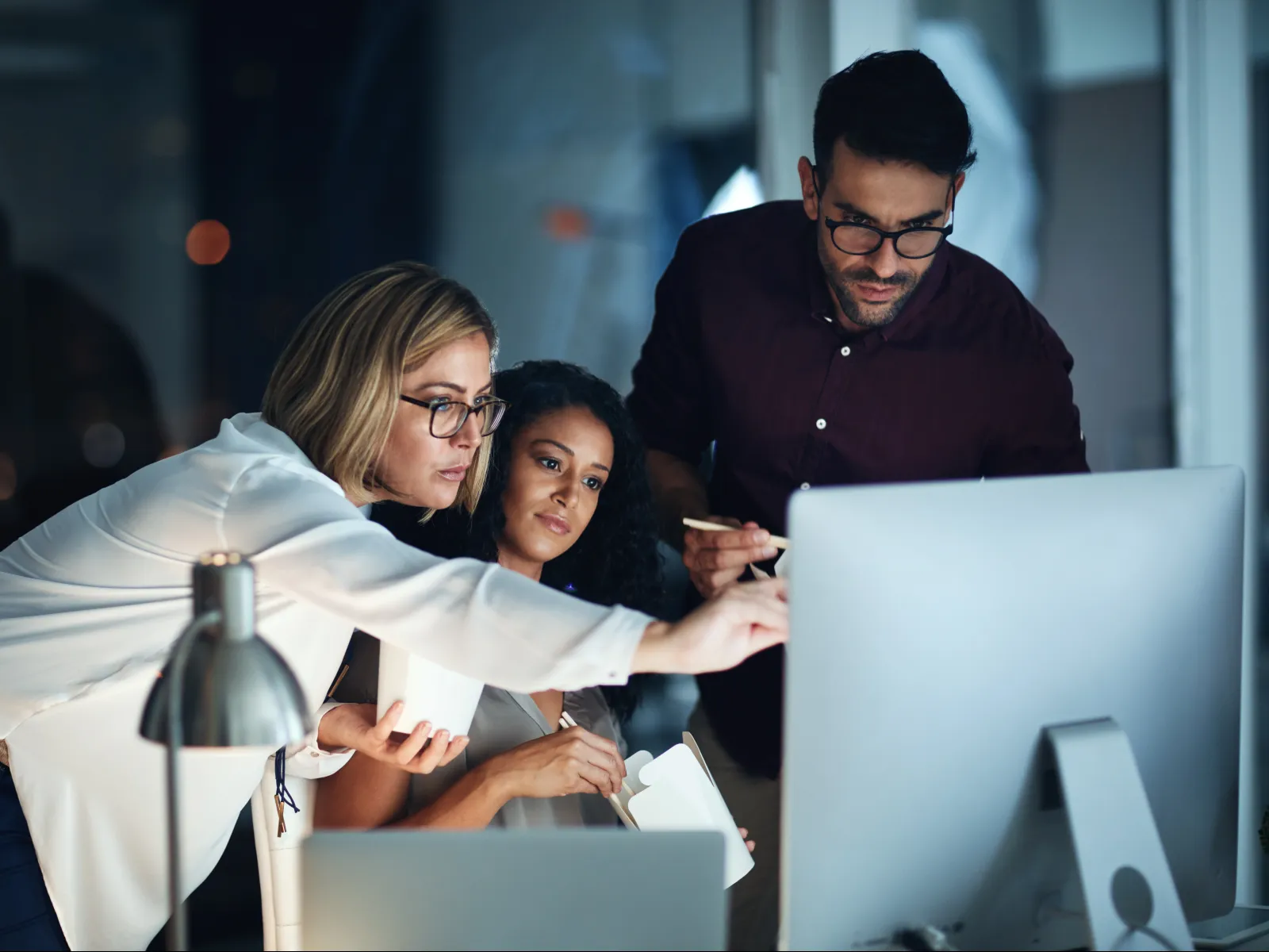 Three professionals collaborating over a computer in an office during a late-night work session.