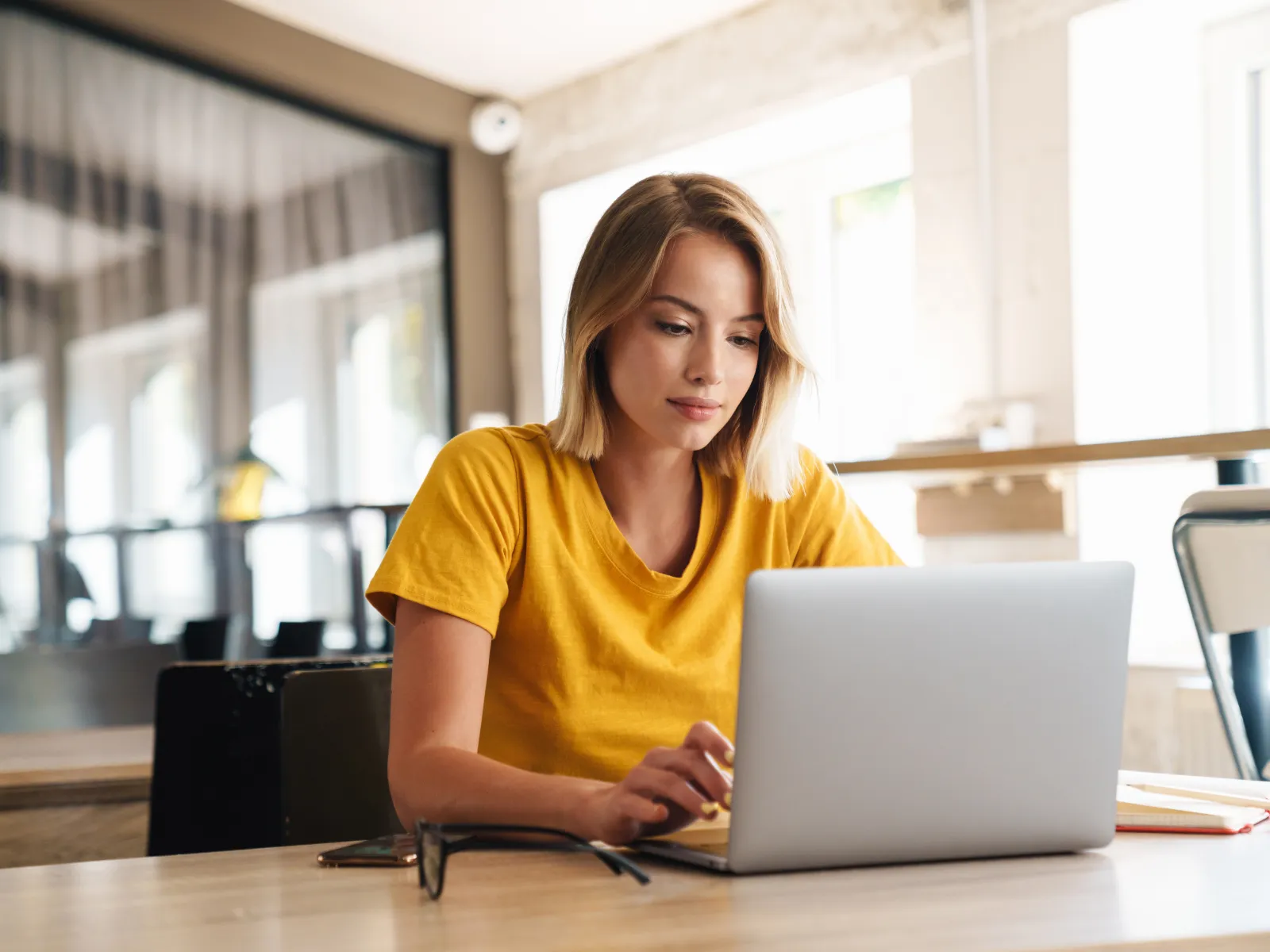 Young woman in yellow shirt working on a laptop in a modern cafe setting.