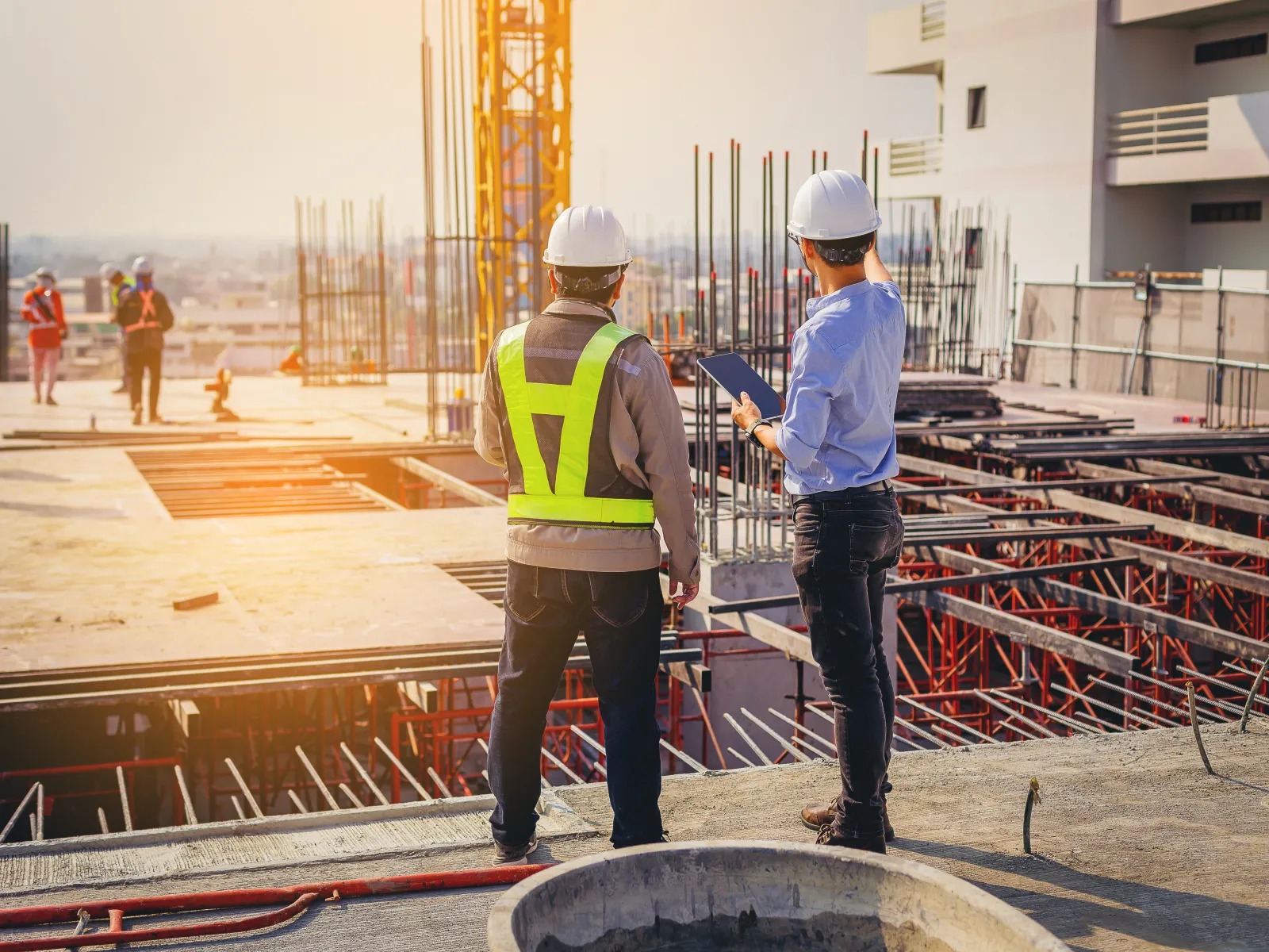Construction workers discussing plans on a building site during sunset with safety gear and machinery.