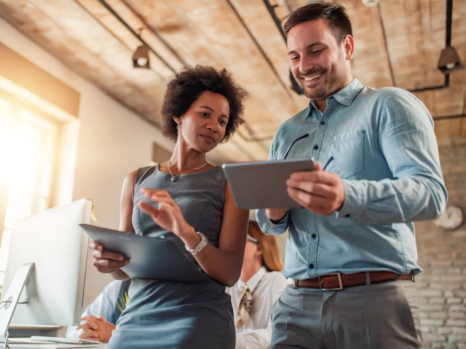 Two professionals collaborate in a modern office, discussing data on tablets and laptops.