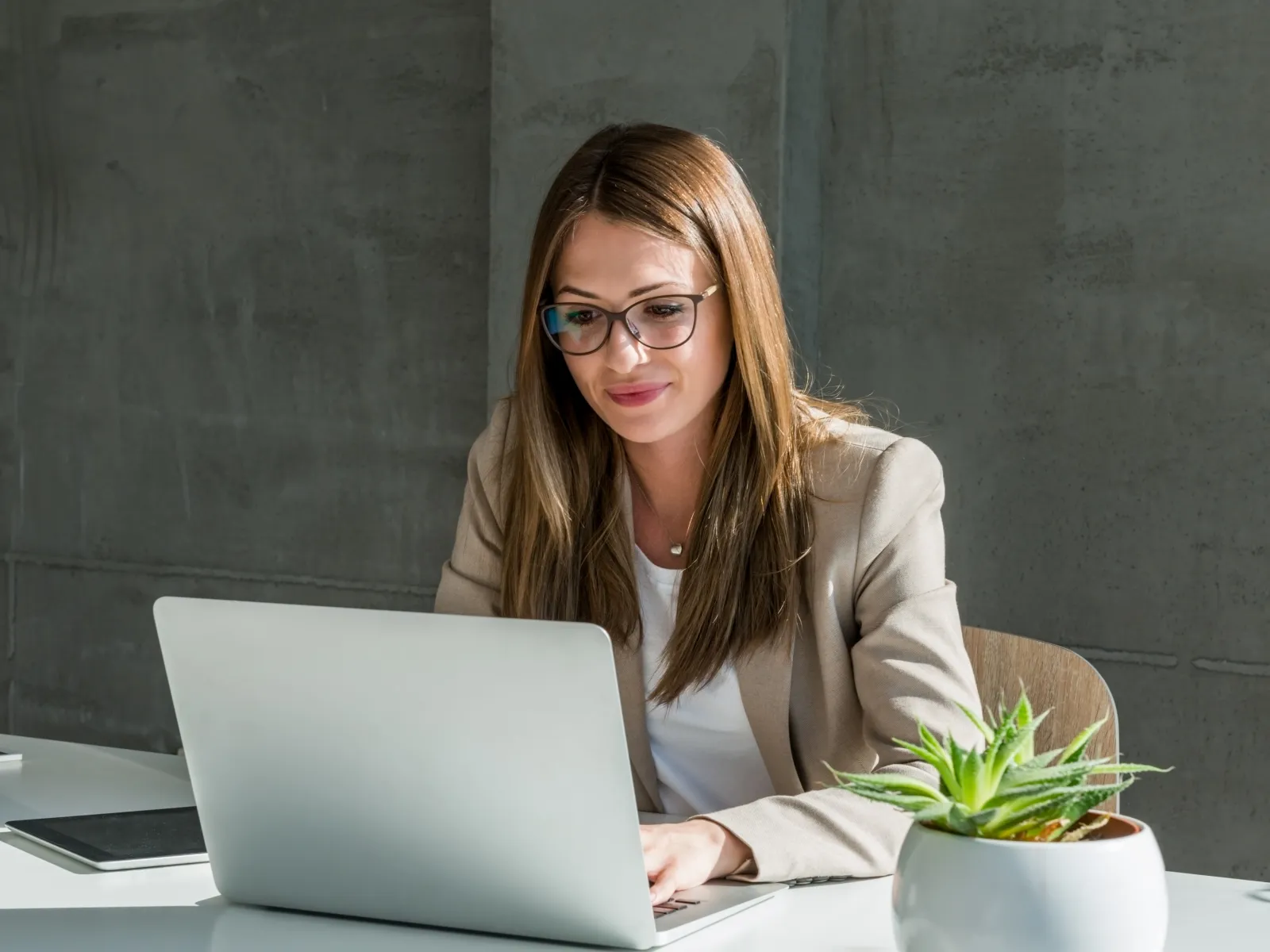 A professional woman working on a laptop in a modern office setting with plants and a coffee cup.