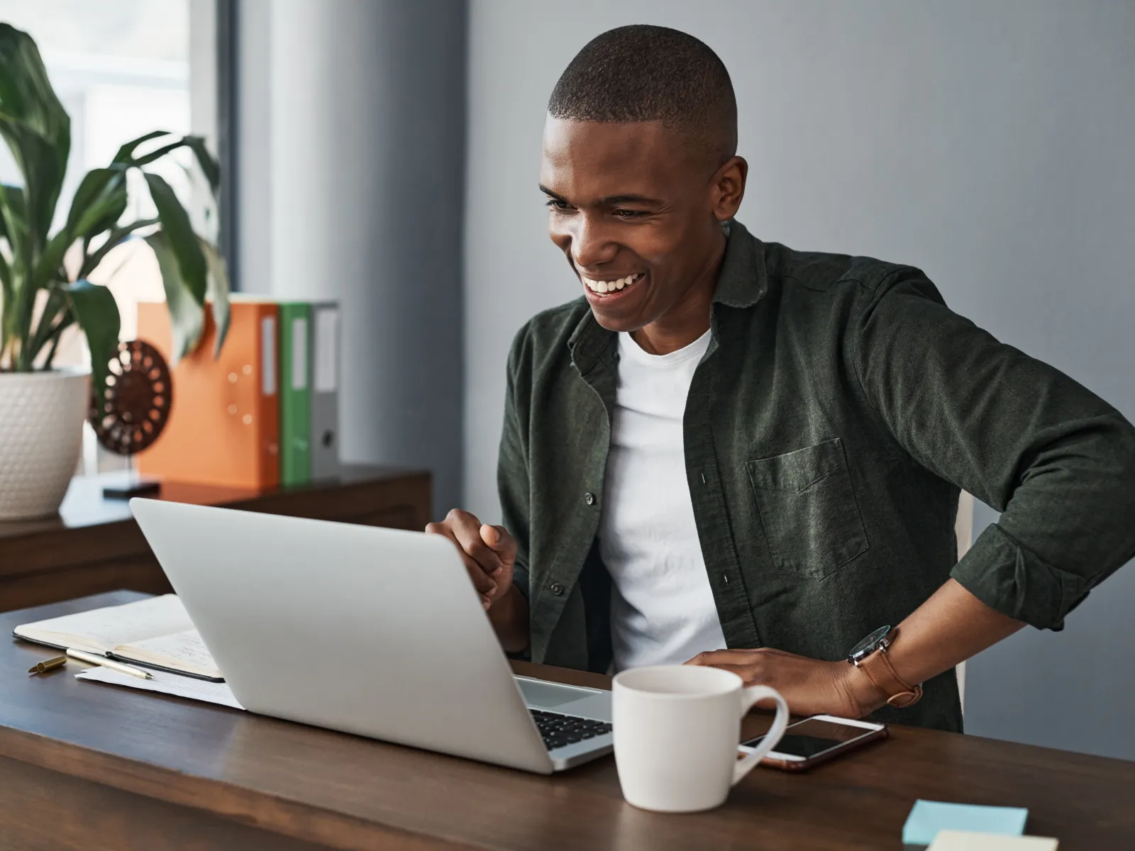 Smiling man using a laptop at a desk with a plant and stationery in a bright workspace.