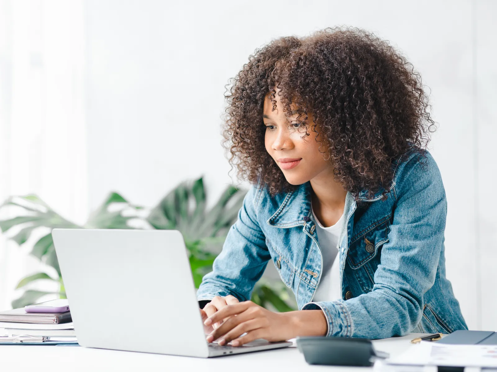 Woman with curly hair working on a laptop at a bright desk with plants around her.